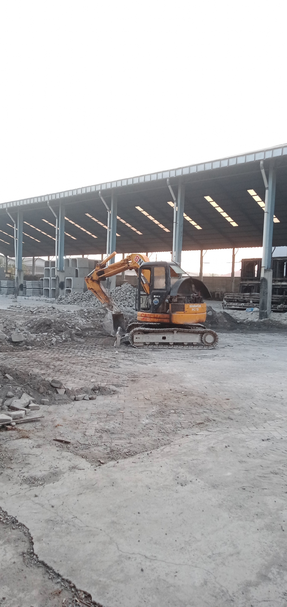 The image shows a yellow Komatsu PC75 excavator in an outdoor industrial setting in Sumput, Indonesia. The ground is a mix of concrete and dirt, littered with concrete debris. Behind the excavator, there is a pile of concrete blocks and building materials. A large open-sided structure with a corrugated metal roof supported by steel beams is in the background. The sky is overcast. There are no people visible in the image.