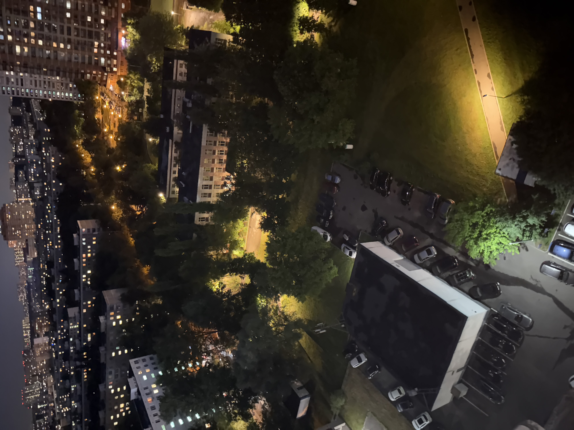 This is a high-angle, aerial nighttime photograph of a residential area in Moscow, Russia. The scene depicts a dense urban environment with numerous multi-story apartment buildings, illuminated by lights from windows and streetlights. Trees are interspersed throughout the cityscape, some casting shadows. In the foreground, there is a flat-roofed building, possibly a commercial or administrative structure, with several cars parked in front of and around it. A parking lot is visible to the left of this building, also with several parked vehicles. A pathway with a lamppost is illuminated by a bright yellow light in the grassy area to the right. The overall impression is one of a typical urban neighborhood at night, with the city lights creating a warm glow against the dark sky.