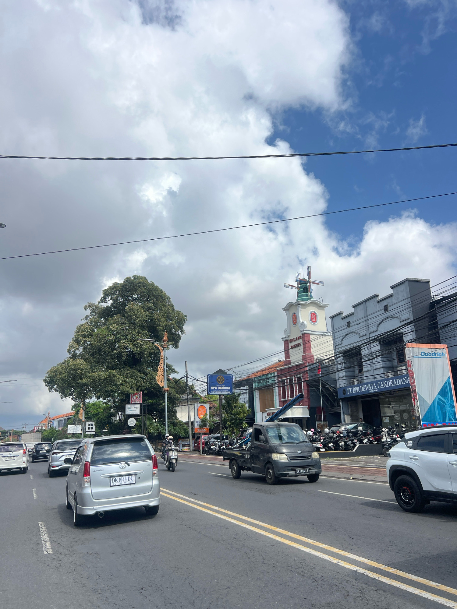 The image depicts a street scene in Denpasar, Indonesia, with a moderate amount of traffic under a partly cloudy sky. In the foreground, a silver Toyota Avanza with the license plate "DK 1044 DC" is traveling in the right lane of a two-lane road. To its left, other vehicles, including a white SUV and a black pickup truck, are also visible. A large, leafy tree dominates the left side of the frame, partially obscuring the buildings behind it.
Further down the road, a distinctive building with a red and white facade and a windmill-like structure on top stands out. The building appears to house commercial establishments, with signage visible, including "HOLLAND BAKERY". Adjacent to it, a grey building with a modern facade displays the text "PT. BPR DEWATA CANDRADANA". A billboard for "Goodrich" is also present.
Several motorcycles are parked along the sidewalk in front of the buildings. Pedestrians are not clearly discernible, but a person on a motorcycle is visible in the middle ground. Overhead, power lines crisscross the sky, with a prominent black wire running horizontally across the upper portion of the image. The road surface is asphalt, marked with yellow double lines. The overall atmosphere suggests a typical day in a bustling urban environment.