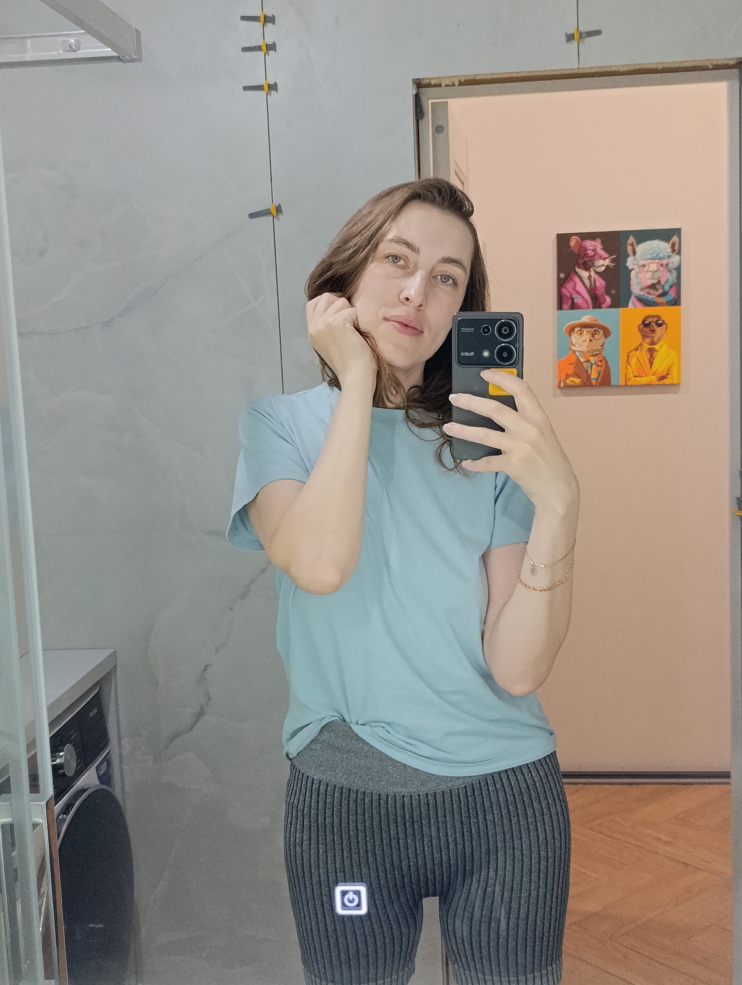 A woman is taking a selfie in what appears to be a bathroom. She is wearing a light blue t-shirt and grey striped shorts. The background includes a mirrored wall, a washing machine to the left, and a door with artwork on it to the right. Tile spacers are visible near the ceiling of the wall with the washing machine. The woman is holding a smartphone in her right hand.
The presence of the washing machine and tiles suggests the bathroom location. The image does not contain explicit information or clues to pinpoint the exact city. The claim of the location being Chaikovsky, Russia is speculative and not discernible from the visual data.
