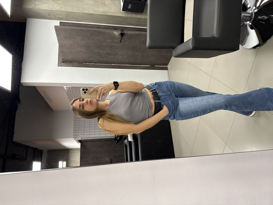 A woman takes a selfie in a mirror inside a modern-looking establishment, potentially a salon or shop. She wears a grey tank top, blue jeans with a black belt, and a smartwatch. She holds a smartphone in her right hand. Behind her, a hair styling chair is visible along with a dark gray door. The setting has a tiled wall section and neutral-toned flooring.