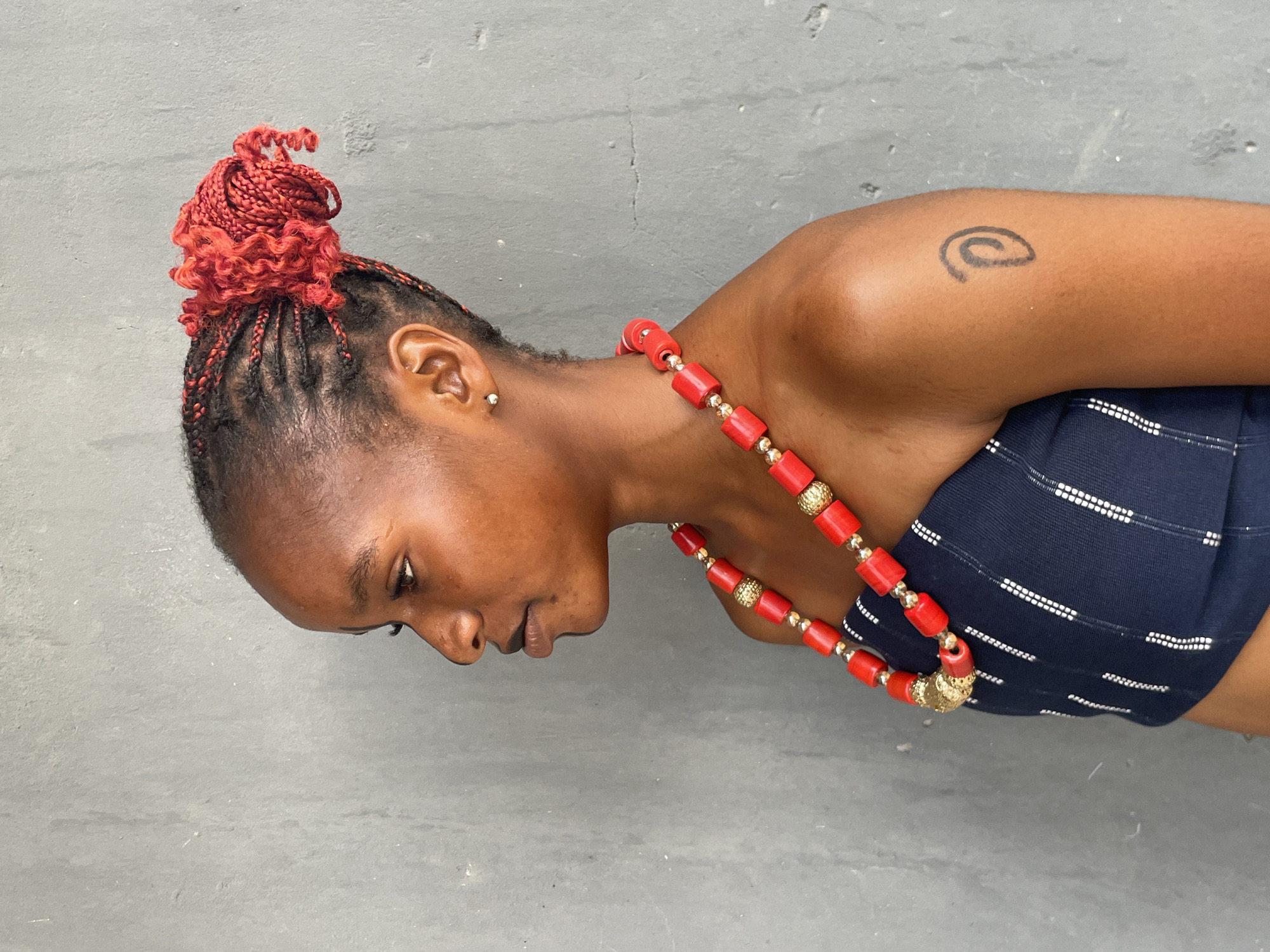 A side profile portrait captures a person with a dark skin tone, likely in Badagry, Nigeria. The individual wears a striking red and gold beaded necklace and a dark blue top with thin white horizontal and dashed patterns. Their hair is styled in tightly woven red braids, with dark roots visible, gathered into an elaborate, textured bun on top of their head. A small, simple silver stud earring is visible in their right ear.
The person is looking downwards, with a calm or pensive expression. A distinct black spiral tattoo is visible on their upper right arm. The background is a plain, light grey wall, which appears to be a textured concrete surface, providing a neutral backdrop that highlights the subject. There is no discernible activity other than posing for the photograph. No text is visible in the image. The lighting is soft and even, suggesting an overcast day or an indoor setting.