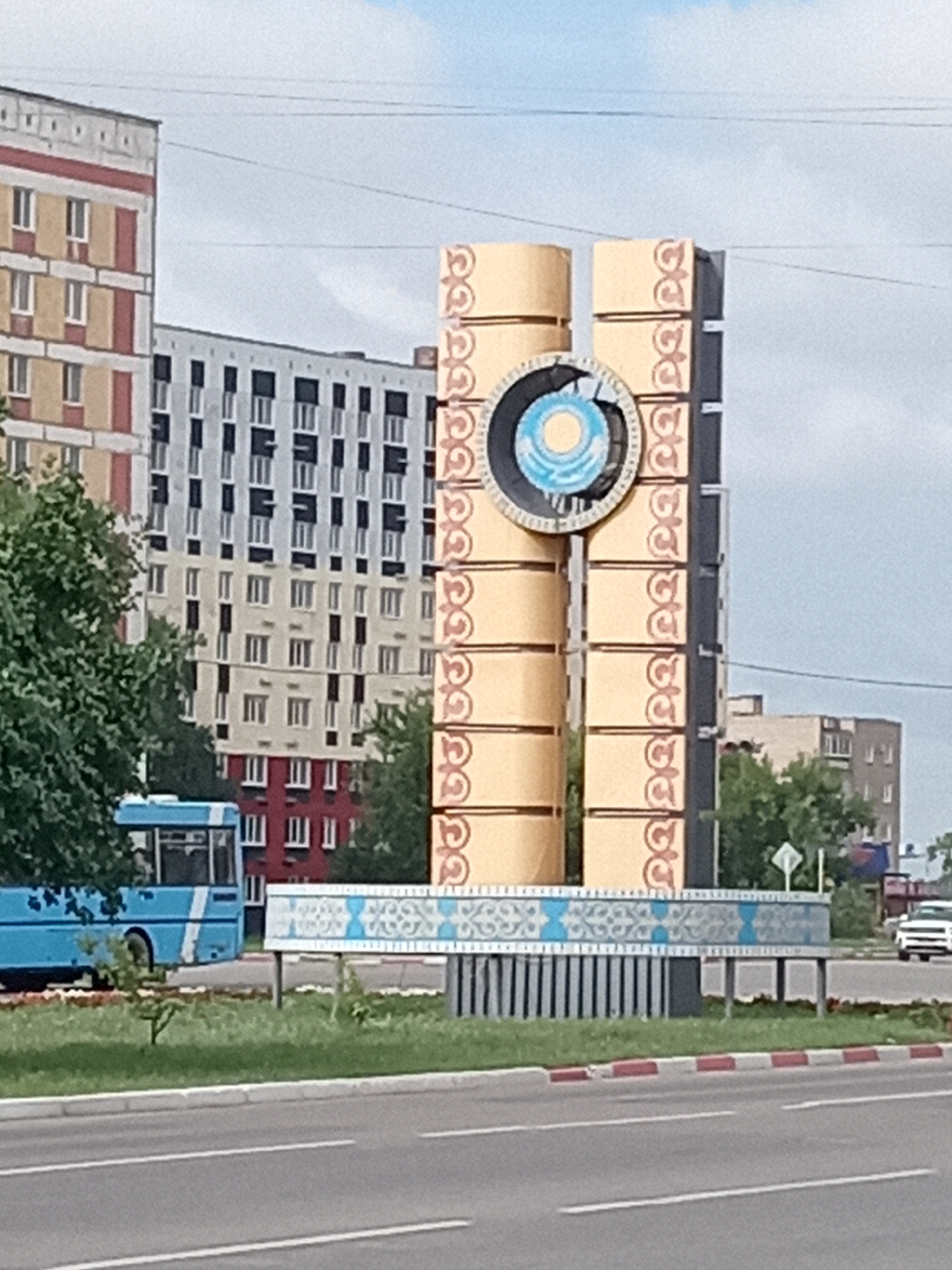 A street-level view captures a monument in Rudny, Kazakhstan. The monument is a tall, abstract structure composed of several stacked rectangular blocks with ornate detailing. At its center, a circular emblem featuring a sun and a stylized pattern is visible. The monument is situated in a public space with a road in the foreground and several multi-story buildings in the background, including an apartment building with a red base and upper floors of cream and red. A blue bus is parked on the left side of the image, and a white car is visible on the right side of the road. Green grass and trees line the area around the monument. The sky is overcast with some visible clouds.
