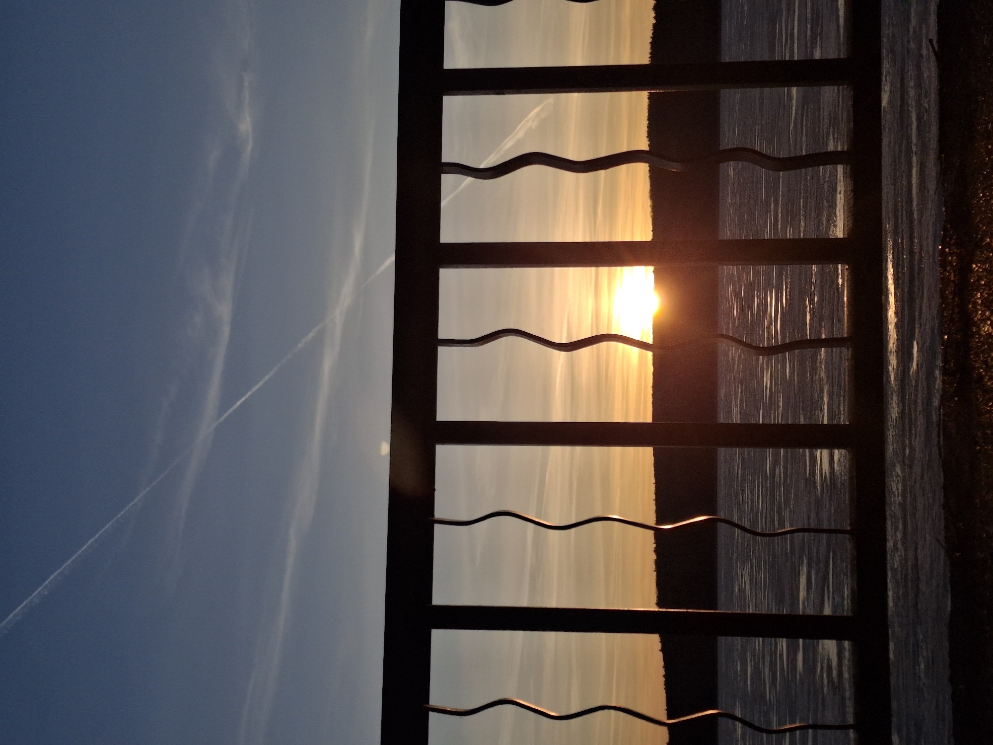 The image captures a serene outdoor scene during either sunrise or sunset, overlooking a body of water, likely the Kama Reservoir, in Chaikovsky, Russia. The scene is divided horizontally by a silhouetted fence, with the sky above and the water below.
The sky exhibits a dramatic gradient of colors, transitioning from a deep, clear blue on the left to soft pastels of light blue, white, and warm yellow and orange towards the right horizon. Wispy cirrus clouds are scattered across the sky, along with prominent white contrails from an aircraft, cutting diagonal lines through the blue expanse.
The sun, a brilliant and intense golden-orange orb, is positioned on the right side of the frame, partially obscured by the dark structure of the fence. Its radiant light creates a powerful glow.
A dark, possibly metallic, fence or railing spans horizontally across the middle ground. It features sturdy vertical posts and multiple horizontal bars, with distinctive undulating (wavy) decorative elements running between the main horizontal supports. This structure is heavily silhouetted against the bright light, appearing almost black.
Below the fence, the vast expanse of water stretches into the distance. The surface vividly reflects the warm light of the sun and sky in elongated, vertical streaks of gold and orange, interspersed with darker, rippled areas that reflect the deeper blues of the sky. The far right edge of the image reveals a dark, somewhat textured shoreline or bank.
The time of day is undoubtedly the golden hour, either sunrise or sunset, evident from the low position of the sun and the warm, ethereal glow bathing the landscape. The weather appears clear, with only high-altitude clouds and contrails present. No people are visible in the tranquil scene, and there is no discernible text.