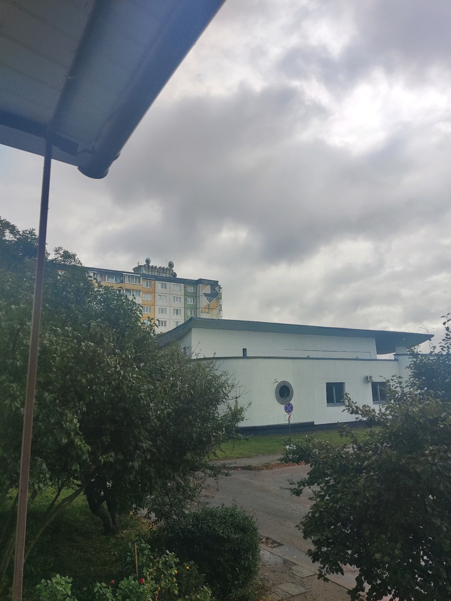 This image captures an overcast sky above a multi-story apartment building and a lower, white-walled building with a dark roof. The apartment building features a facade with sections of yellow, white, and green, and has numerous windows and what appear to be satellite dishes or ventilation units on its roof. The white-walled building has a distinctive circular window and two rectangular windows, along with an air conditioning unit. In the foreground, lush green trees with dark leaves partially obscure the buildings. A dark pole, possibly part of a balcony railing or support structure, is visible on the left side of the image. A paved road or walkway with scattered leaves is visible between the trees and the white-walled building. A traffic sign with a red circle and a blue background with a white cross is situated near the circular window. The overall scene suggests an outdoor view in an urban or suburban environment under cloudy weather conditions.