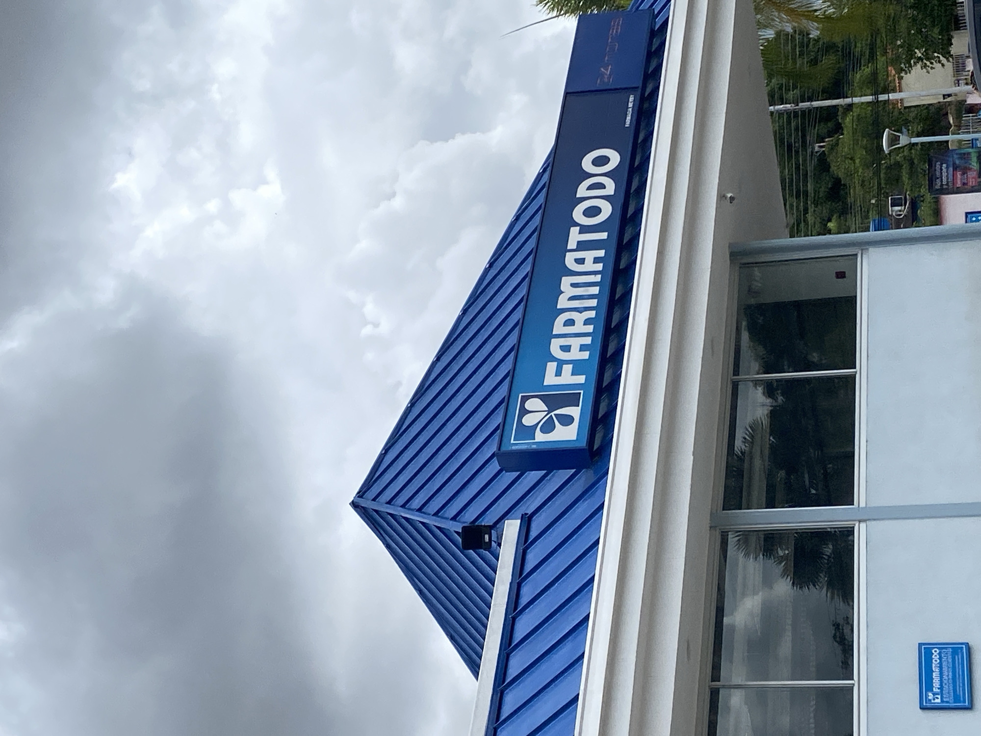 The image shows a Farmatodo pharmacy in Tinaquillo, Venezuela. The blue sign with white lettering reads "Farmatodo" vertically. The pharmacy has a blue roof and white exterior walls with windows reflecting the sky. A smaller sign with the Farmatodo logo is visible on the wall near the window. Overcast sky with clouds are in the background. Some green foliage can be seen in the background as well.