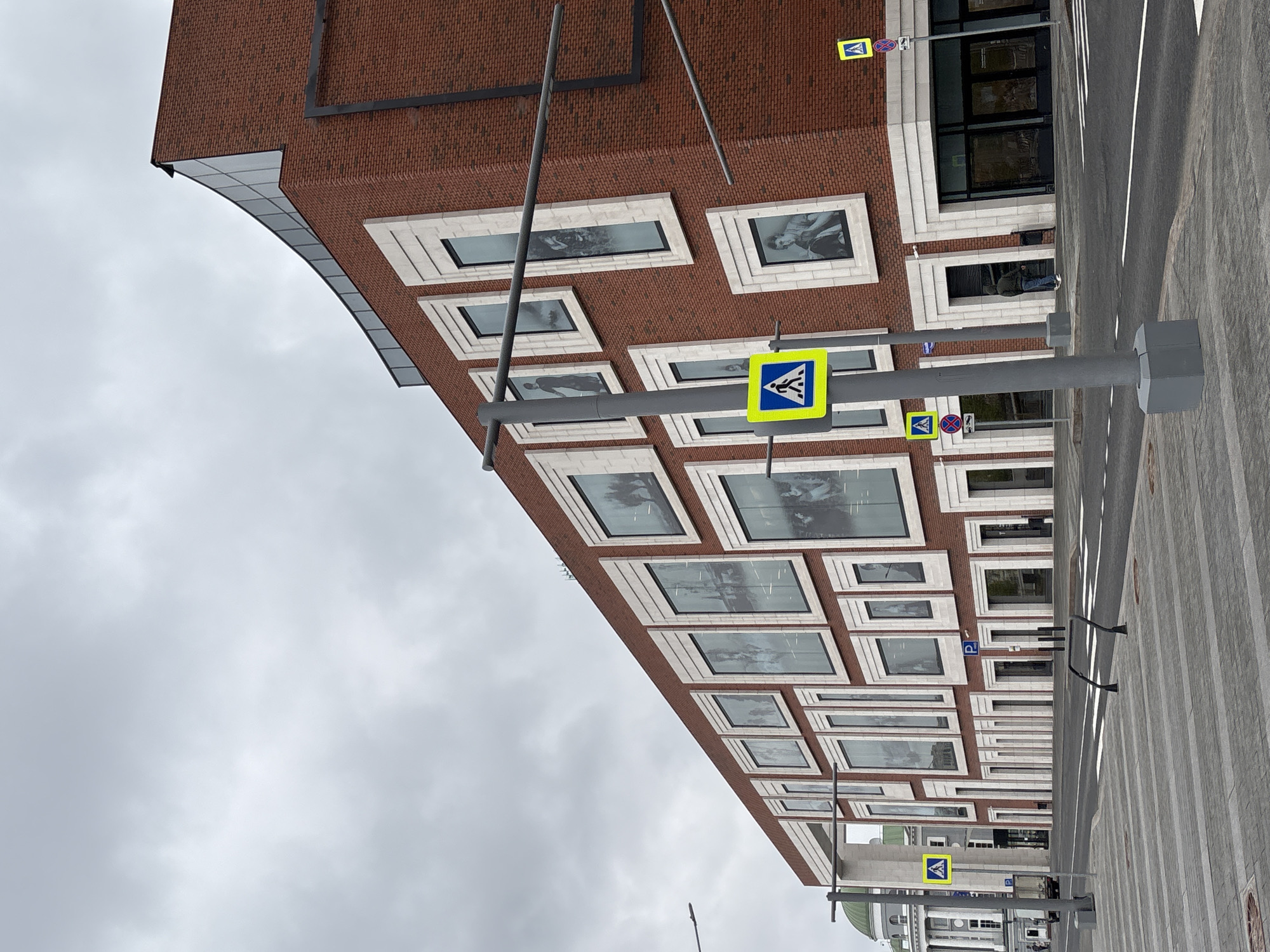 The image depicts a portion of the State Tretyakov Gallery on Krymsky Val in Moscow, Russia. The building is constructed of red brick with white stone window frames. Pedestrian crosswalk signs are visible along the sidewalk in front of the gallery. Images or exhibits can be seen in the windows. The sky is overcast.