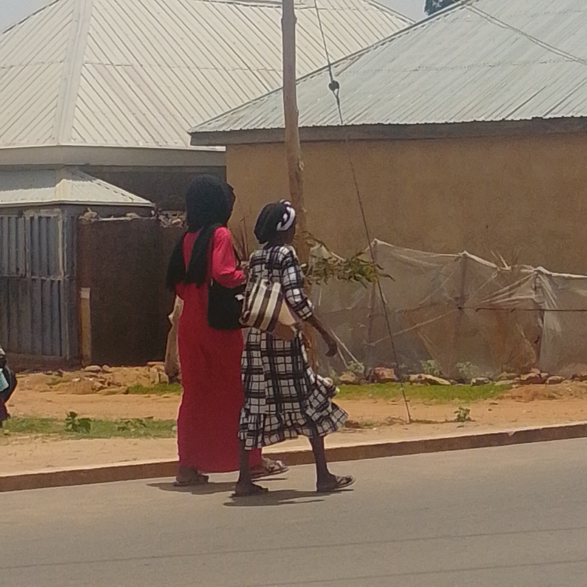 Two individuals are walking on a paved road in Gombe, Nigeria. The person on the left is wearing a long red garment and a black head covering, carrying a black bag over their shoulder. The person on the right is wearing a black and white checkered dress and a black head covering with a white stripe, also carrying a black and white striped bag. Behind them, there are buildings with corrugated metal roofs and walls made of mud or concrete, and a fence made of corrugated metal. The ground appears to be a mix of dirt, rocks, and sparse vegetation. The scene is outdoors during daylight.