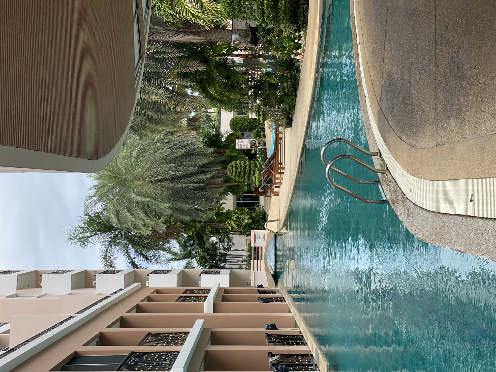 The image shows a long, narrow swimming pool adjacent to a multi-story building with a beige facade. The pool has turquoise water and is bordered by a textured concrete walkway. Metal handrails emerge from the water near the foreground. In the background, a small arched bridge crosses the pool, connecting two sides of the building complex. Palm trees and lush green vegetation are visible on the other side of the pool, suggesting a tropical or subtropical environment. The sky is overcast. The overall scene depicts an outdoor amenity area of a hotel or condominium in Pattaya City, Thailand, characterized by its resort-like atmosphere. There are no people visible in the image.