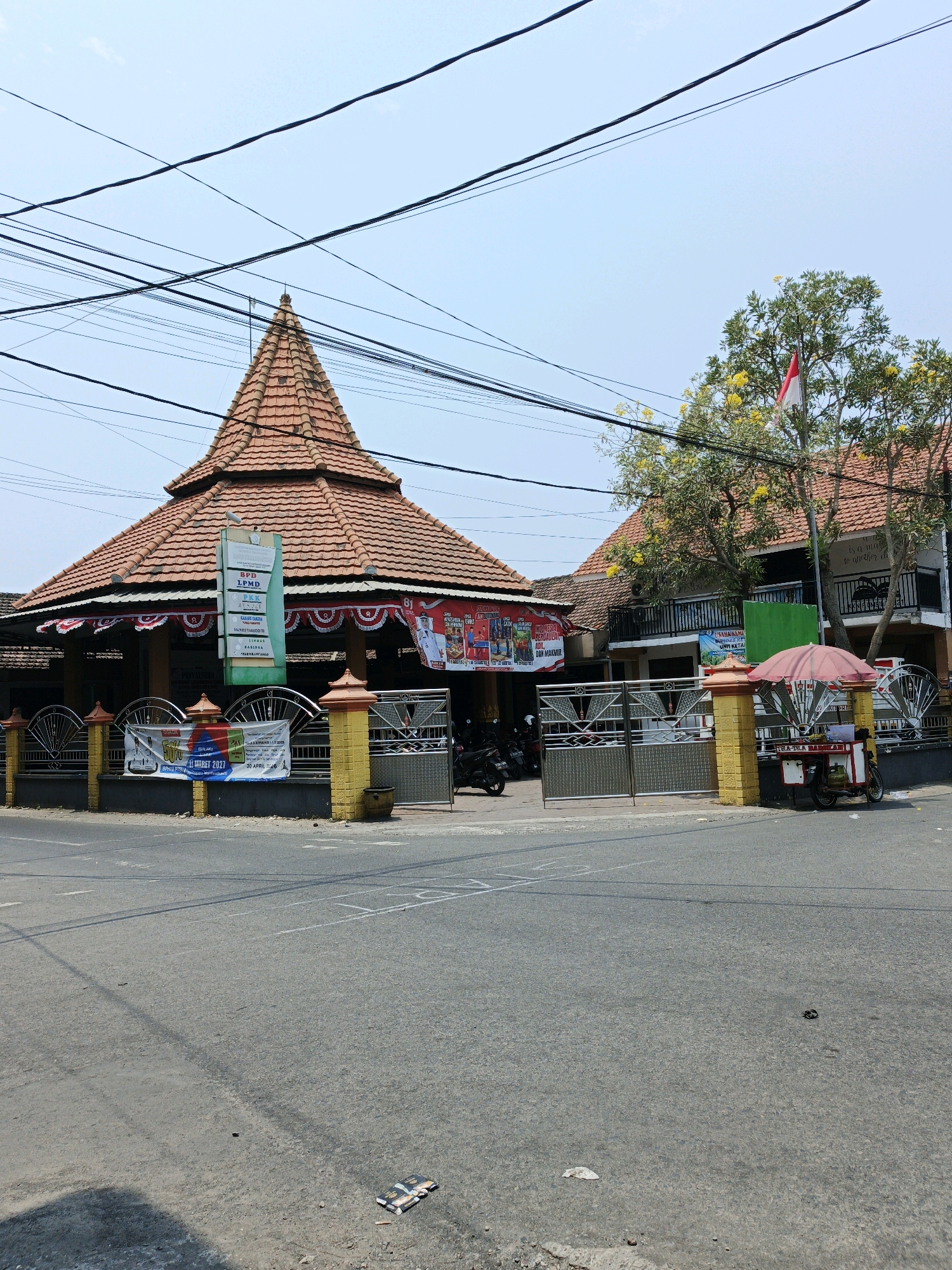 The image depicts a street scene in Ketegan, Indonesia, on a clear day. In the foreground, a paved road is visible with some white markings. To the right of the road, a street vendor's cart with a pink umbrella is parked. The cart has a small gas tank and appears to be selling food or drinks. Behind the cart, a gated entrance leads to a building with a red-tiled, multi-tiered roof. The building's facade is decorated with red and white banners and a flag. A large tree with yellow flowers stands next to the building, with an Indonesian flag planted on its branches. A number of power lines crisscross the sky above the scene. The overall impression is of a lively, urban environment, likely a local market or public space, given the presence of the vendor and the festive decorations.