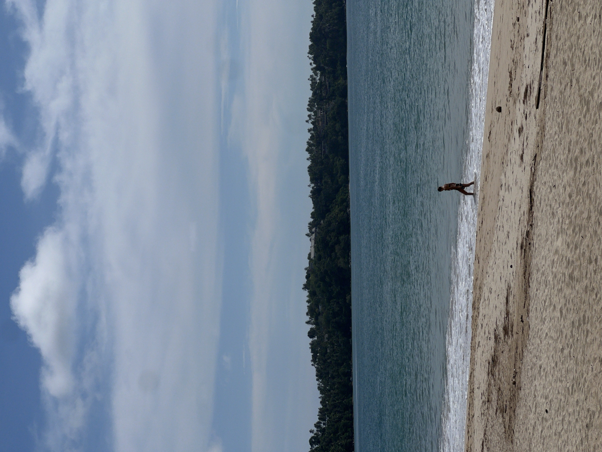 The image shows a beach scene. A person is walking along the shoreline where the water meets the sand. In the background, there is a landmass covered with dense greenery. The sky above is filled with clouds.