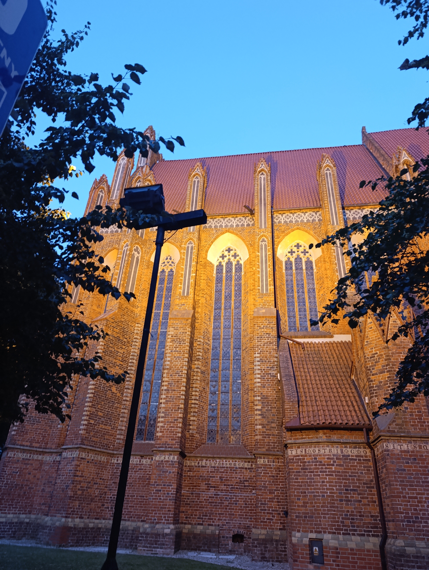 The image shows the exterior of the Cathedral Basilica of St. John the Baptist and St. John the Evangelist in Toruń, Poland, during twilight. The cathedral is constructed of red brick and features Gothic architectural elements, including pointed arched windows with intricate tracery and a tiled roof. The building is illuminated by warm, artificial lighting, highlighting its textured brickwork and decorative details.
In the foreground, a dark silhouette of a streetlamp with a crossbar is visible on the left side of the image. Dark green foliage from trees partially obscures the view of the cathedral on the left and right edges of the frame. The sky above is a clear, deep blue, indicative of the evening hours. There are no people or animals visible in the scene. The ground in front of the cathedral appears to be covered in grass, with some dark patches suggesting shadows or dampness. A small, square electrical box with a yellow hazard symbol is mounted on the lower right section of the brick wall. The overall impression is one of architectural grandeur bathed in the warm glow of evening light.