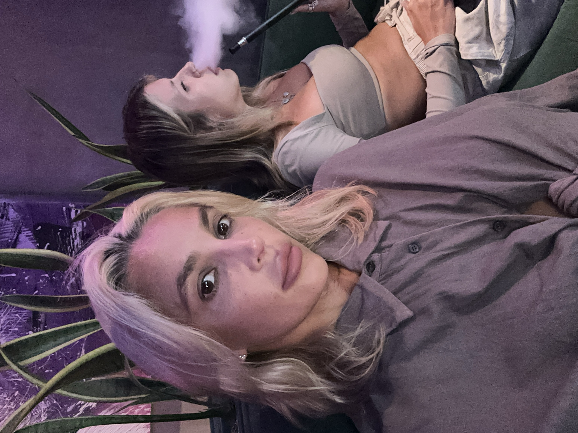 Two women are seated on a green sofa in a dimly lit room, likely a lounge or bar. The woman in the foreground, with blonde hair and fair skin, is looking directly at the camera with a neutral expression. She is wearing a light purple button-down shirt. To her right, the second woman, also with blonde hair, is facing away from the camera, exhaling smoke from a hookah. She is wearing a light-colored crop top and light-colored pants. The background is dark, with a large potted plant with green and yellow leaves to the left of the frame. The overall lighting is dim, with purple hues cast on the wall and plant. The environment suggests a relaxed, social setting, possibly a shisha lounge, in Kuta Selatan, Indonesia.