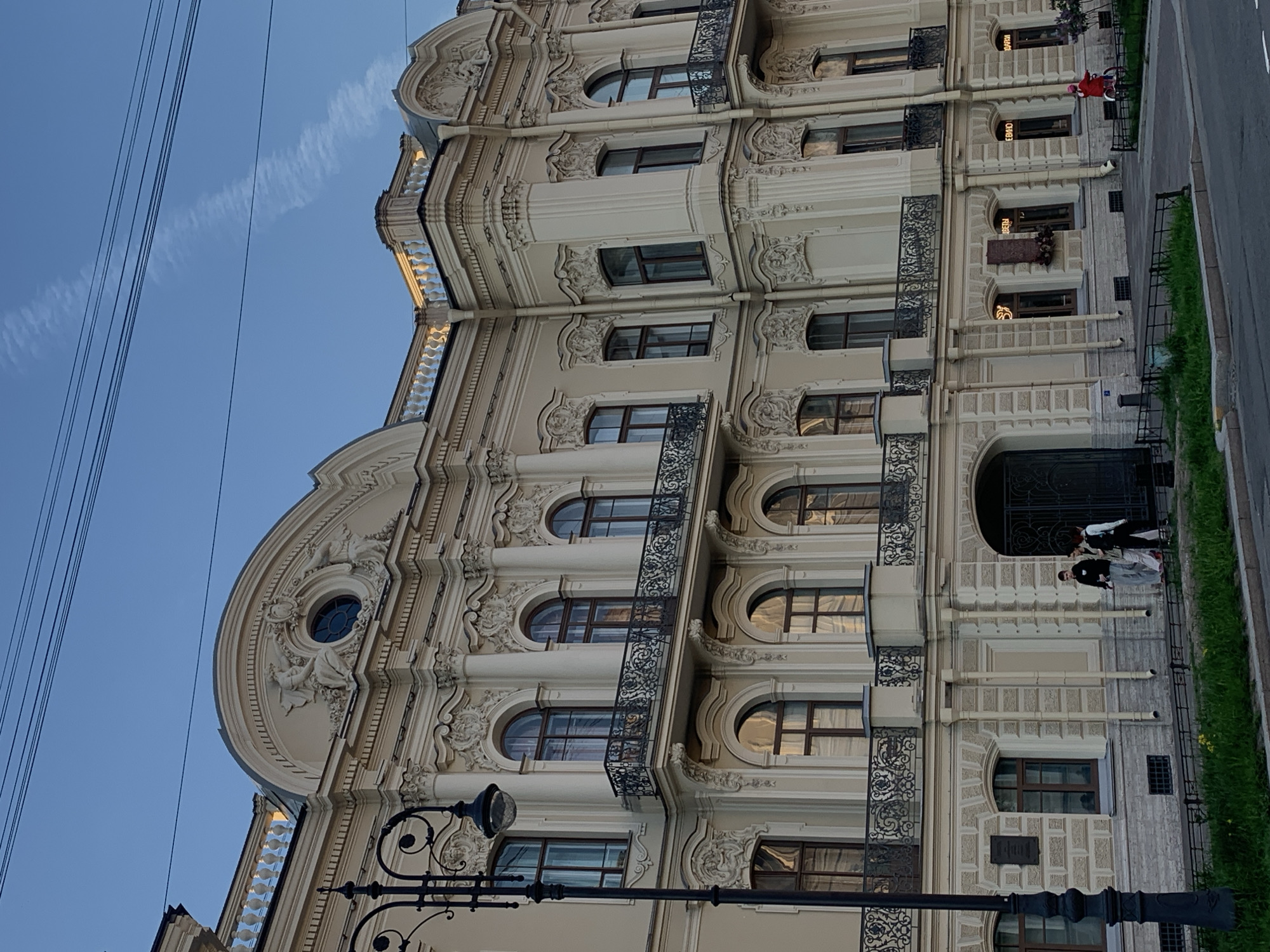 Here's a description of the image:
The image depicts an ornate building facade in Saint Petersburg, Russia. The building features elaborate architectural details, including arched windows, decorative molding, and wrought iron balconies. A round window with sculptural ornamentation is visible on the upper portion of the facade.
A section of street and sidewalk is visible on the right, with grass. A lamppost is in the foreground. A cluster of people are on the landing next to the front door. Power lines are also present in the sky to the left of the building.