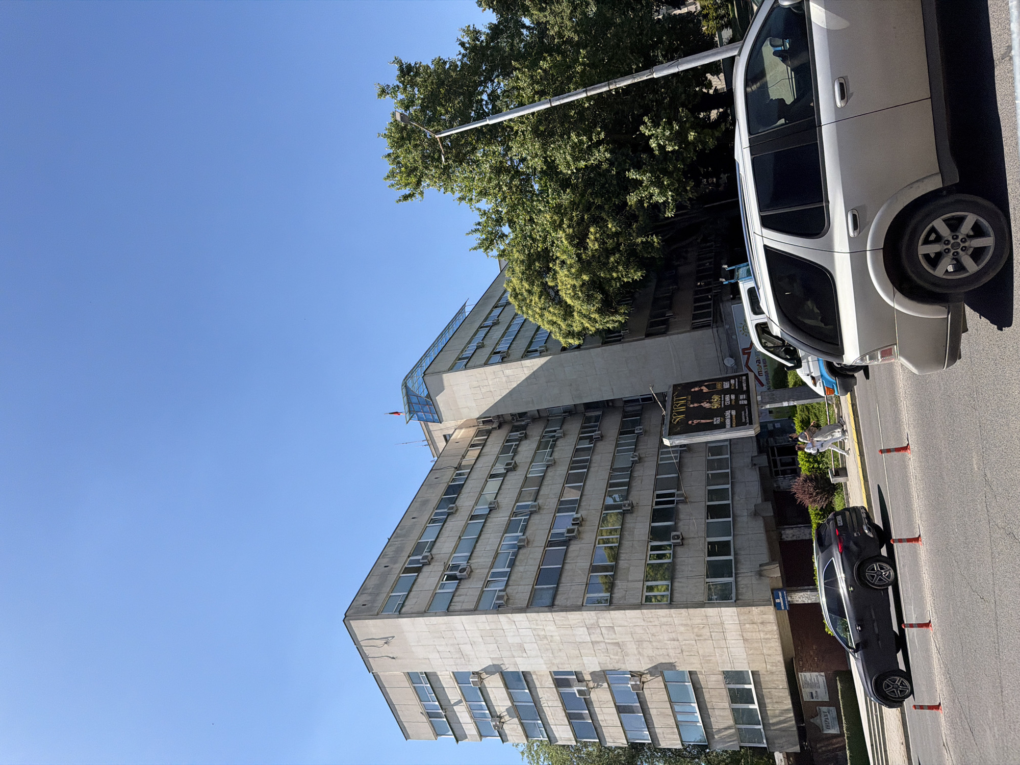 The image depicts a street scene in Plovdiv, Bulgaria, dominated by a large, multi-story building with numerous windows and air conditioning units. The building has a modern design with a prominent glass section on the upper part. A tree is situated beside the building. A white minivan is parked on the right side of the image, taking up significant space. A dark grey car is parked on the street as well, near orange traffic cones. Two individuals are seen sitting on a bench near the building, though details about them are obscured. A commercial sign is located on the side of the building near the street. The sky is clear and blue.