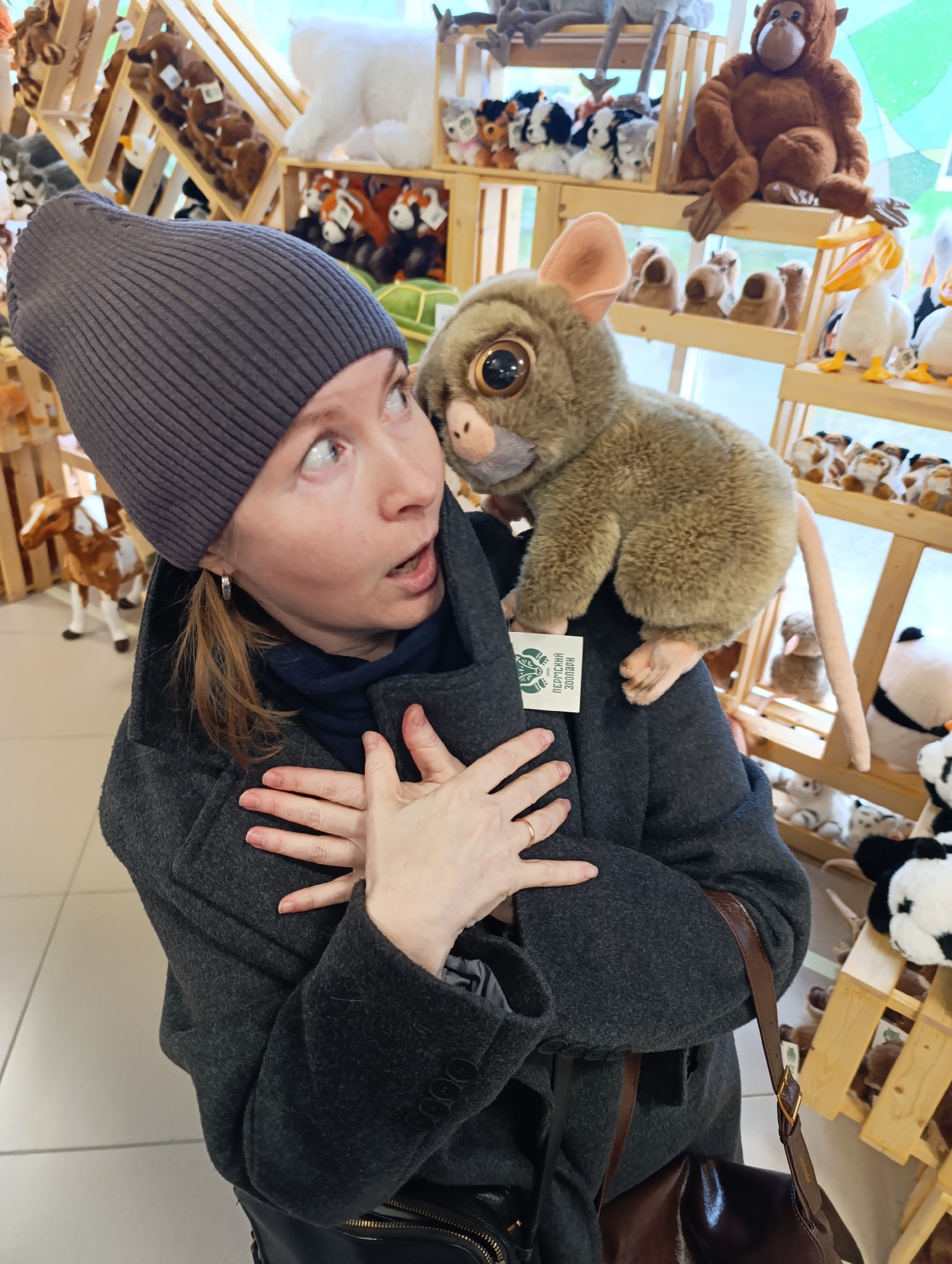 A fair-skinned woman wearing a gray wool coat and a gray knitted hat is posing with a plush tarsier toy perched on her shoulder in a store that sells stuffed animals. The woman's hands are clasped near her chest, and she is looking to the left with a surprised expression. Shelves filled with various stuffed animals, including bears, pandas, pelicans, and monkeys, are visible in the background. The store is located in Perm, Russia, as indicated by the text on the tag attached to the tarsier. The floor has a light-colored tile pattern. The woman also has a dark-colored bag hanging from her shoulder.