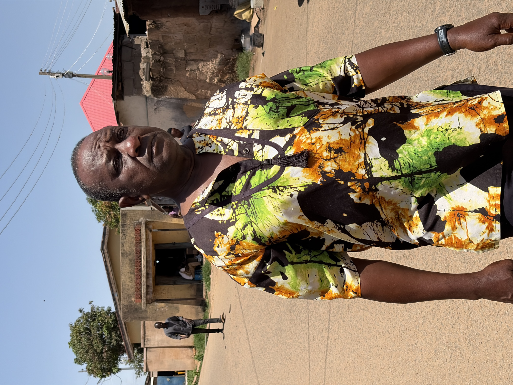 A portrait-style photograph captures a middle-aged man standing outdoors, looking directly at the camera with a neutral expression. He has a dark complexion and is wearing a short-sleeved, button-up shirt with a vibrant, abstract pattern in black, green, yellow, and white, featuring a black tassel detail at the collar. A black wristwatch with "LACOSTE" faintly visible is on his right wrist.
The man stands on a light-colored, possibly unpaved, ground surface under a clear blue sky. The bright sunlight suggests it is midday or early afternoon. In the background, several structures are visible, consistent with a town or village setting in Jos, Nigeria. To the left, a building with a red corrugated iron roof and stone-like walls is seen, along with a wooden utility pole and multiple power lines. Another person is partially visible near this building. To the right, a lighter-colored building with a small portico and an open doorway stands, with another individual visible further down the street. Sparse green foliage is also present in the background.