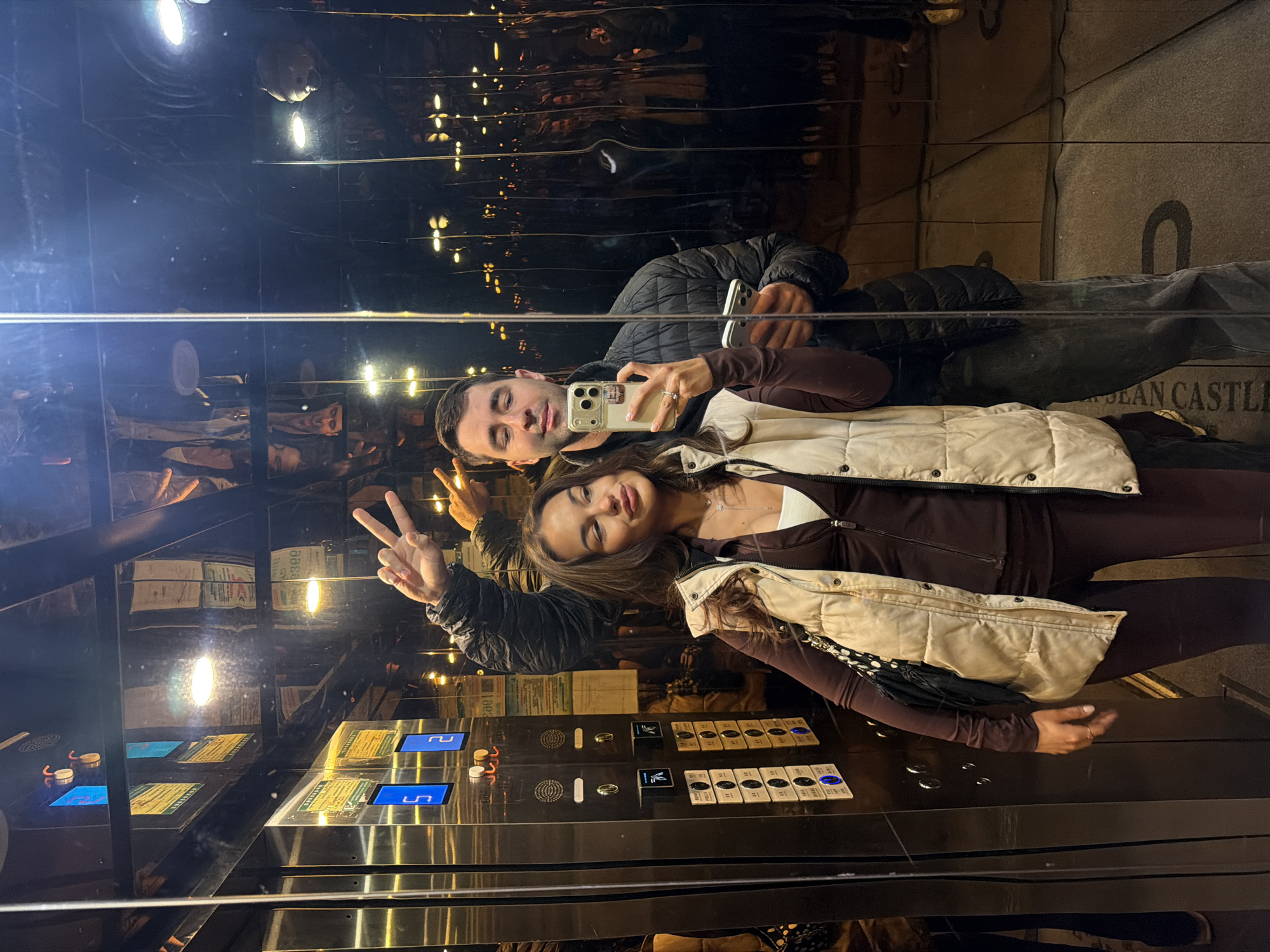 A couple is taking a selfie in an elevator, likely in Yongding District, China. The man, wearing a black puffer jacket, is holding the phone. The woman, wearing a brown outfit with a cream-colored vest, is making a peace sign. The elevator has mirrored walls, creating multiple reflections. The elevator panel with buttons is visible. The background shows an exterior scene at night with lights.