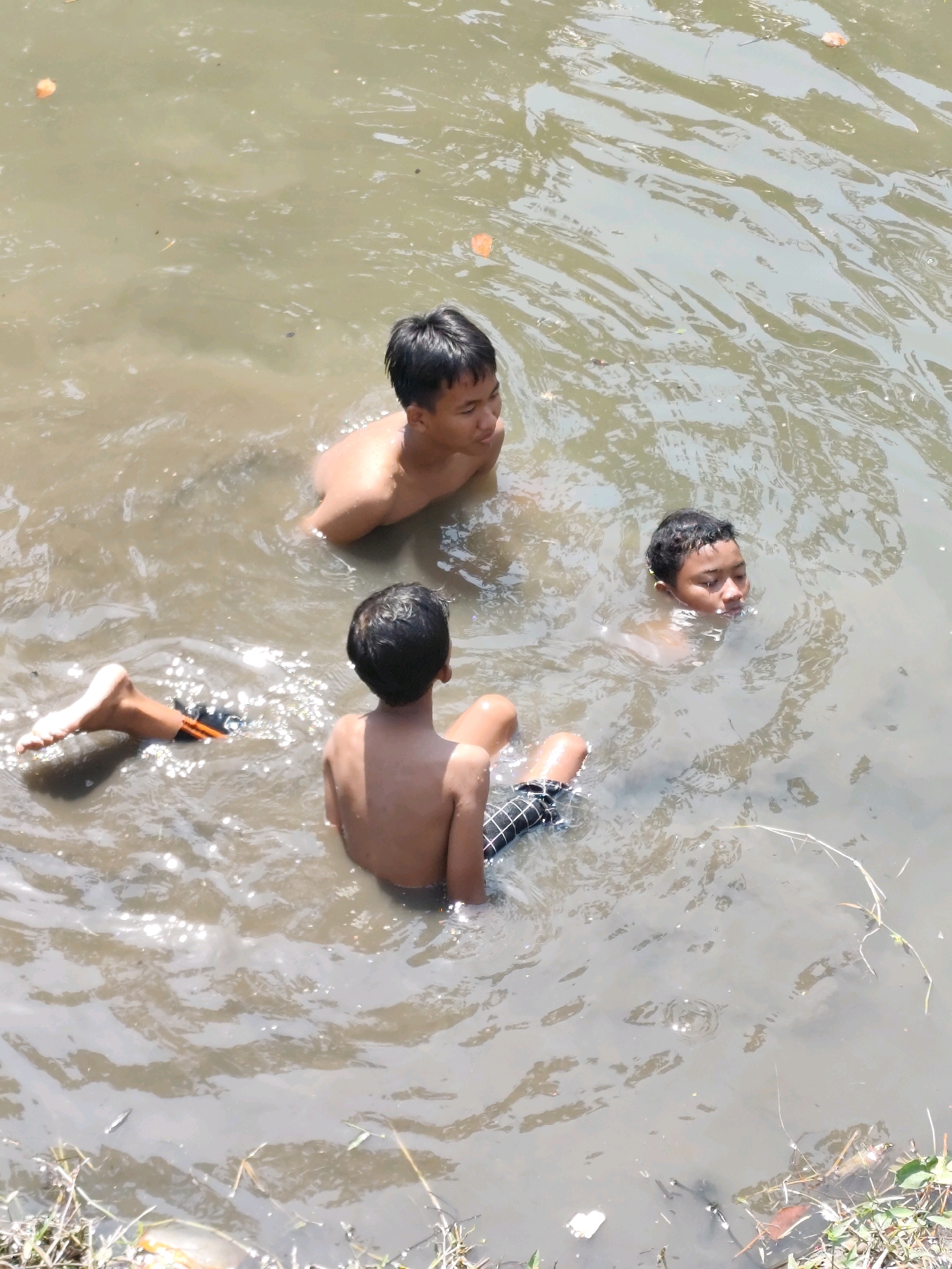 Three young boys are swimming in a shallow, murky body of water under a bright sun. The water is brown with some visible debris and reflects sunlight in ripples. One boy, facing away from the camera, sits in the water with his back to the viewer. His legs are submerged, and he is wearing dark shorts with a checkered pattern. Another boy, facing the camera and looking to his right, is partially submerged with his upper body exposed. He has dark hair and is looking out of frame. The third boy, also facing the camera, has his head and shoulders above the water, looking down. His face is visible, and he has short, dark hair. One of his legs is visible at the edge of the frame, extended into the water. The location is identified as the Bambanglipuro, Indonesia area.
