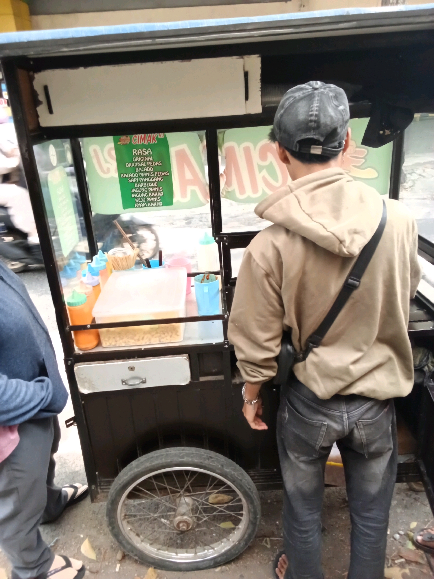 A man wearing a brown hooded sweatshirt and dark jeans stands with his back to the camera, looking at a food cart. He has a black baseball cap on his head and a black crossbody bag slung over his right shoulder. The food cart is black and has a display window showing various food items and condiments. A green sign with text in Indonesian is visible inside the cart. The cart has a large wheel on the left side. The scene appears to be on a street in Bandung City, Indonesia, with a paved ground and some debris. Another person, partially visible on the left, is wearing a blue jacket and gray pants.
