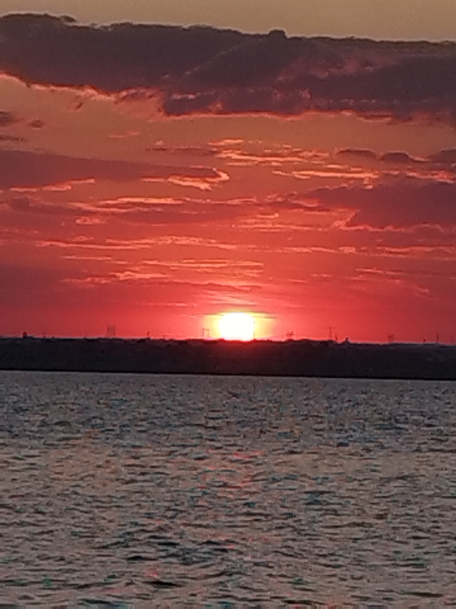 A sunset is captured over the Black Sea in Constanța, Romania. The sky is filled with vibrant red and orange clouds, with the sun appearing as a bright, glowing orb just above the horizon. A dark silhouette of the coastline runs across the middle of the image, punctuated by faint, distant structures that appear to be wind turbines. The water of the Black Sea is visible in the foreground, reflecting the colors of the sky and exhibiting gentle ripples. The scene is devoid of any people or specific identifiable objects on the land or sea.