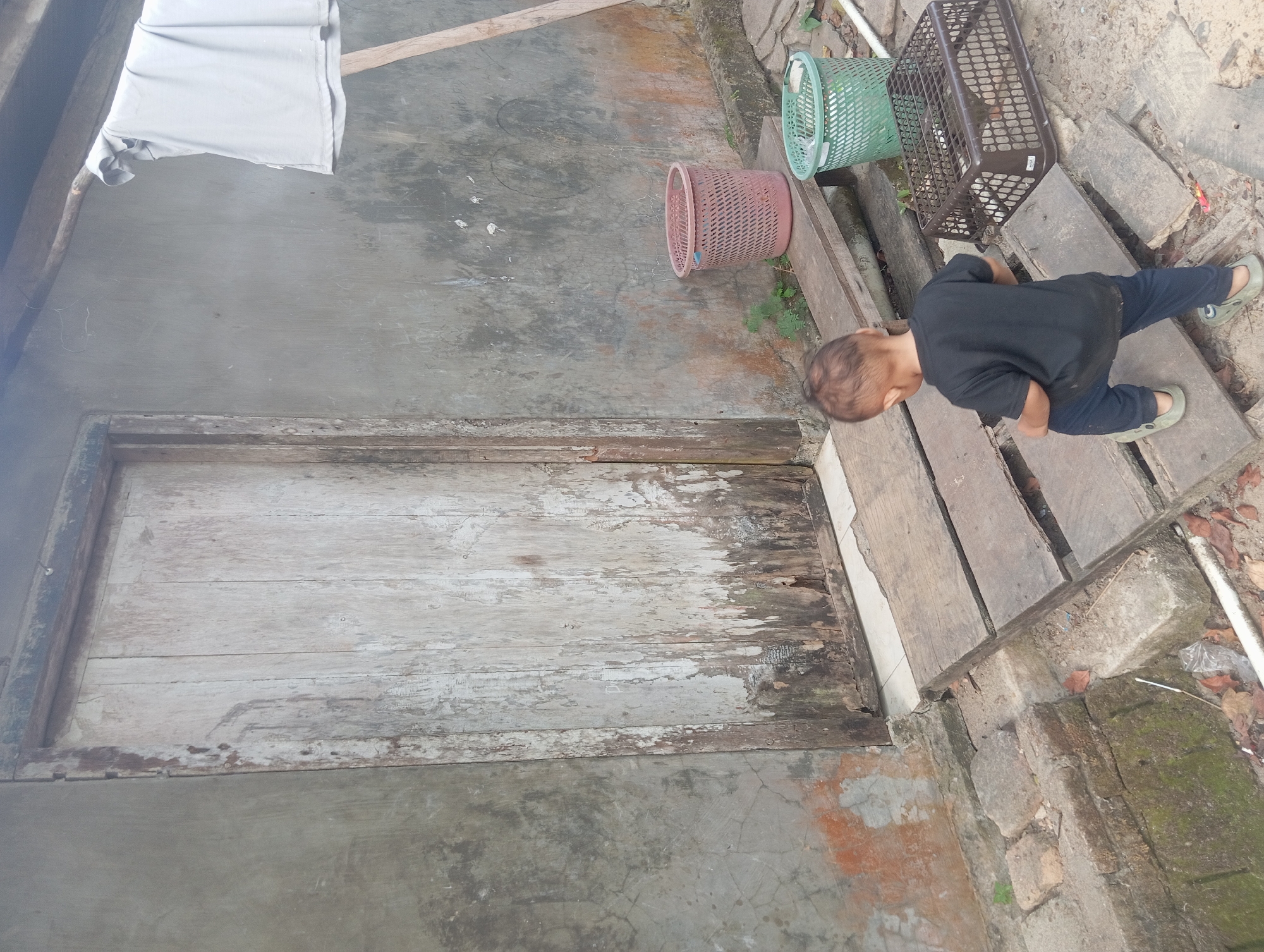 An Indonesian boy, approximately two to three years old, stands on a wooden plank walkway leading to a weathered, light-colored wooden door. He is wearing a black t-shirt and dark blue pants. The boy is facing away from the camera, looking towards the door. To the right of the walkway, there are two plastic baskets, one pink and one green, and a brown plastic crate. The background consists of a rough concrete wall with visible stains and peeling paint, and a wooden beam extending from the wall supporting a gray garment hanging from it. The scene appears to be an outdoor area in Dente Teladas, Indonesia, characterized by rustic construction and an unkempt environment. The ground is covered with dirt, leaves, and some debris.
