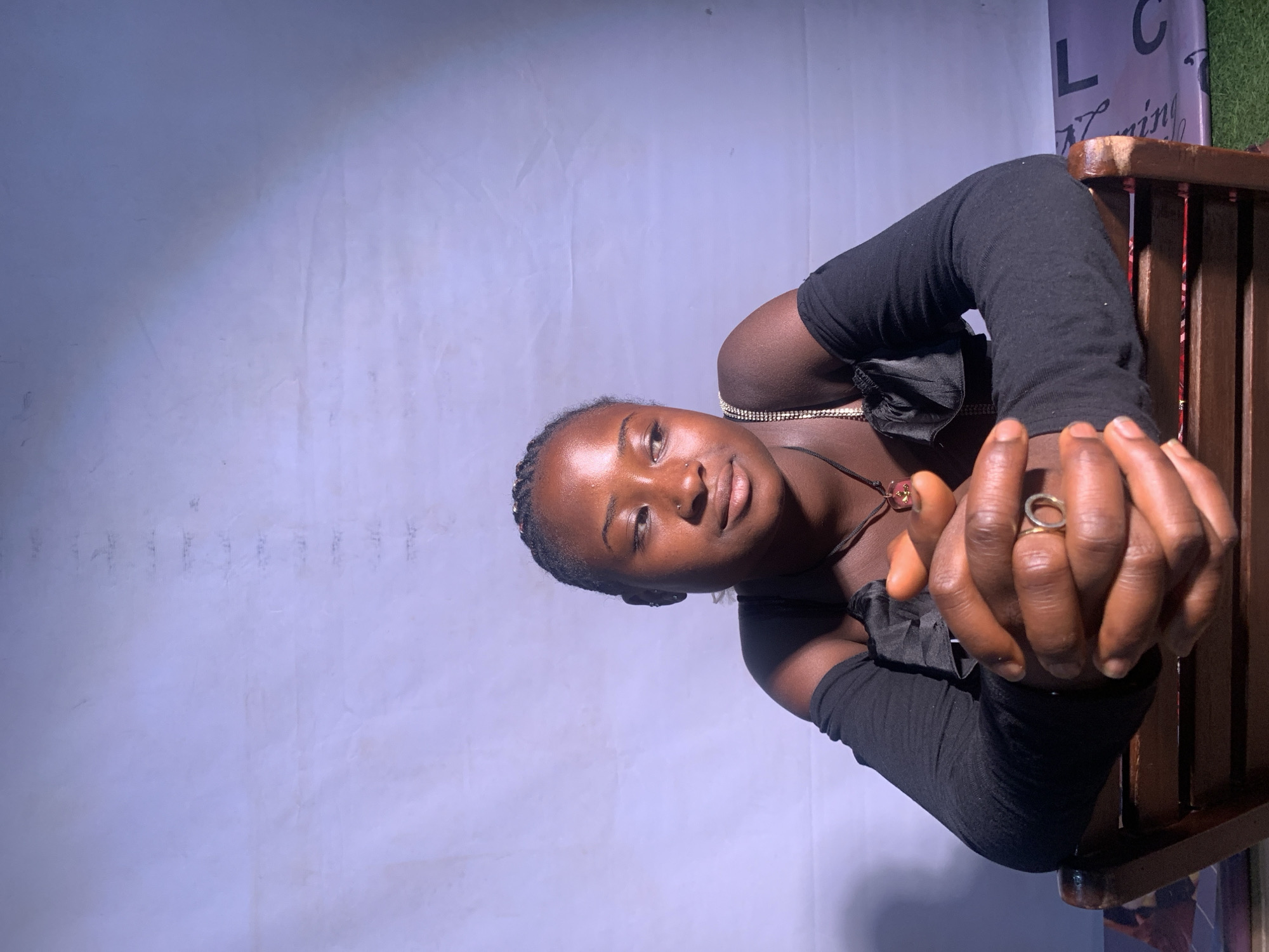 A portrait of a dark-skinned woman taken in a studio setting is depicted. The woman is seated with her hands clasped in front of her, wearing a black top with black arm coverings. She has braided hair and wears jewelry, including a necklace with a pendant and a ring on her finger. She is looking directly at the camera. The background appears to be a studio backdrop with some signage visible on the right side. The setting is likely in Jos, Nigeria, given the prompt's specification.