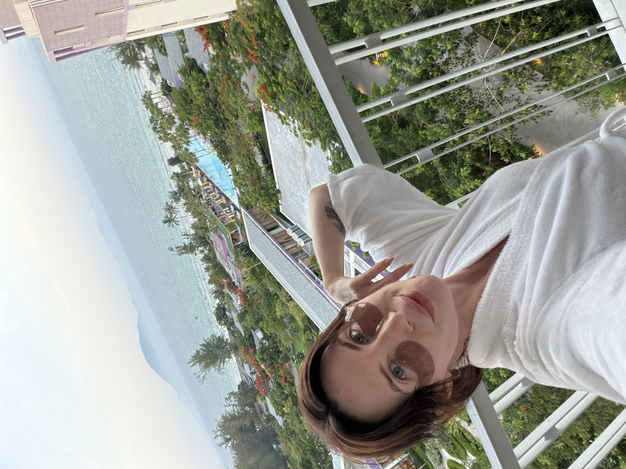 The image presents a high-angle view from a balcony, capturing a woman taking a selfie with a resort and coastline as the background, set in Dong Ninh Hoa Ward, Vietnam.
In the foreground, a woman with shoulder-length dark brown hair and blue-green eyes gazes directly at the camera with a subtle smile. She is wearing a white terry cloth bathrobe and has dark brown gel patches positioned under both eyes. Her left arm is bent, revealing a tattoo on her upper arm, with her hand gently resting near her head. She is leaning against a white metal balcony railing composed of vertical and horizontal bars.
The midground reveals a sprawling resort complex featuring multiple buildings, some with white and purple striped facades, surrounded by extensive green foliage. Palm trees are abundant, and several trees with bright red flowers are visible. A large, irregularly shaped swimming pool with visible swimmers occupies a significant portion of the resort grounds.
Beyond the resort, the background displays a sandy beach populated with numerous individuals, leading into a turquoise-colored ocean. Several small boats are scattered across the water. A range of mountains, partially obscured by atmospheric haze, stretches along the distant horizon beneath a pale, overcast sky. The overall lighting is soft, suggesting either early morning or late afternoon.