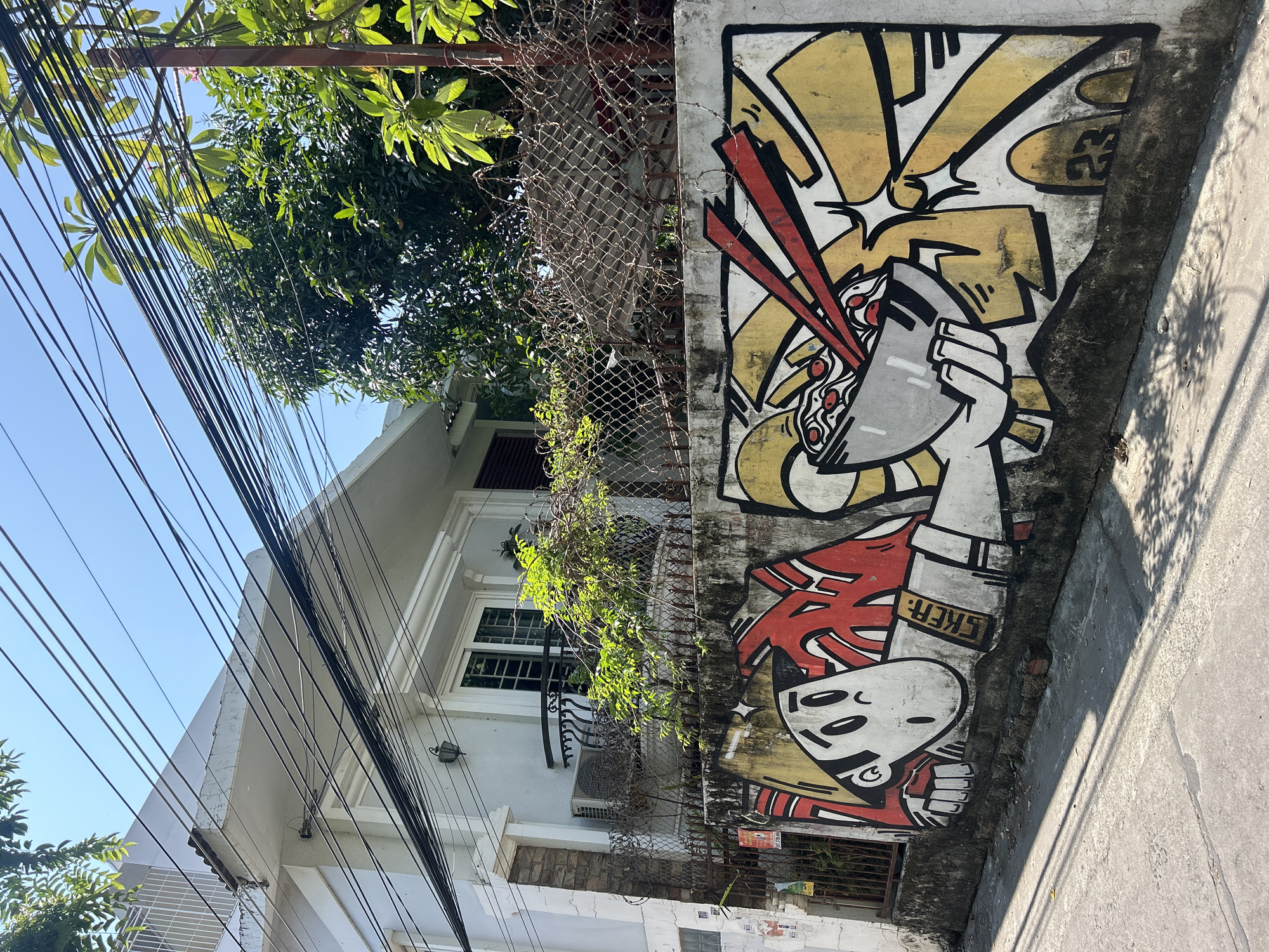 The image presents a street scene in, presumably, Bắc Nha Trang, Vietnam, focusing on a section of a wall adorned with street art. The artwork is rendered in a cartoon-like style, with bold lines and bright colors of yellow, white, and red. The upper portion shows a hand holding a bowl with what appears to be noodles or soup with chopsticks. Below, there is a stylized face with prominent eyes and a red accent. "SKER" is written in gold letters under the face.
Behind the wall, a chain-link fence partially obscures a building. The building is painted a neutral color with white trim and multi-paned windows. Vegetation is growing along the fence, adding a touch of greenery.
A tangle of power lines fills the upper left portion of the frame, contrasting with the clear blue sky visible in the background. The ground is paved with a concrete sidewalk. The light suggests it is daytime, and shadows are cast on the sidewalk.