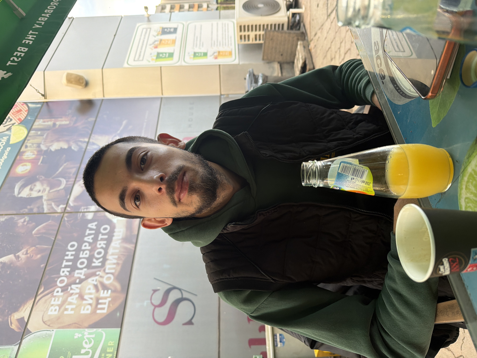 A young man is the central subject of this outdoor portrait, captured in Sofia, Bulgaria, during the daytime. He has short dark hair and a neatly trimmed beard, and is looking directly at the camera with a neutral expression. He is dressed in a dark green hoodie and a black puffer vest.
He is seated at an outdoor table where several items are visible: an open clear glass bottle containing some orange liquid, an empty clear glass bottle, a black paper cup, and a smartphone resting on a stand. The word "ACE" is discernible on the stand. Text on one of the bottles reads "250 ml e".
The background features a building adorned with large advertising posters. One prominent poster displays Cyrillic text that translates to "LIKELY THE BEST BEER YOU WILL TASTE," with parts of the brand name "BER NER" also visible. The word "ulgaria" is partially visible, providing a location cue. Another poster shows a stylized "S" logo. Above, a green awning has the mirrored text "THE BEST" on its underside. An air conditioning unit is also visible on the building facade. The scene is well-lit by natural light, suggesting clear weather.
