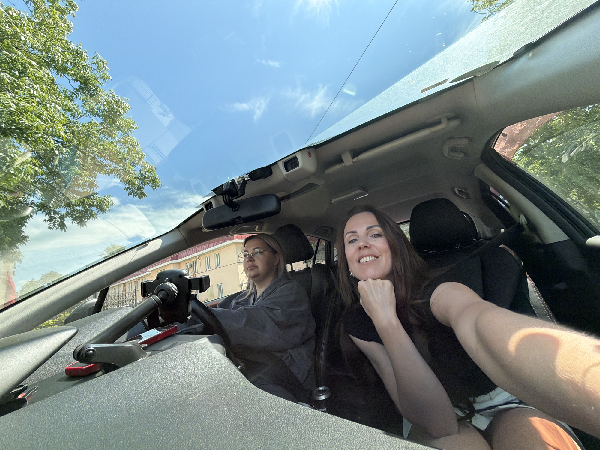 Two women are inside a car. The woman in the driver's seat is wearing glasses and a gray top. The woman in the passenger seat is smiling and taking a selfie. The car has a sunroof and a dashcam. Outside the car, there are trees and a building. The image appears to be taken in Birobidzhan, Russia, based on the user's prompt.