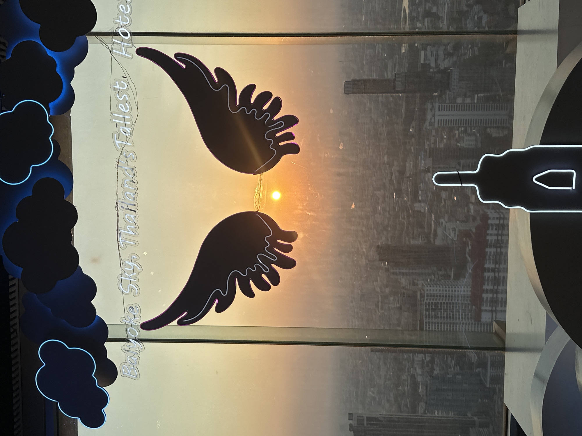 The image depicts a view from the Baiyoke Sky Hotel, Thailand's tallest hotel, in Bangkok. The view is seen through a glass window featuring neon decorations of clouds and angel wings. The sun is setting, visible between the wings, casting a warm orange glow over the cityscape below. The cityscape includes numerous buildings, some of which appear to be reflected in the glass. Another neon decoration in the shape of a bottle is seen to the right.