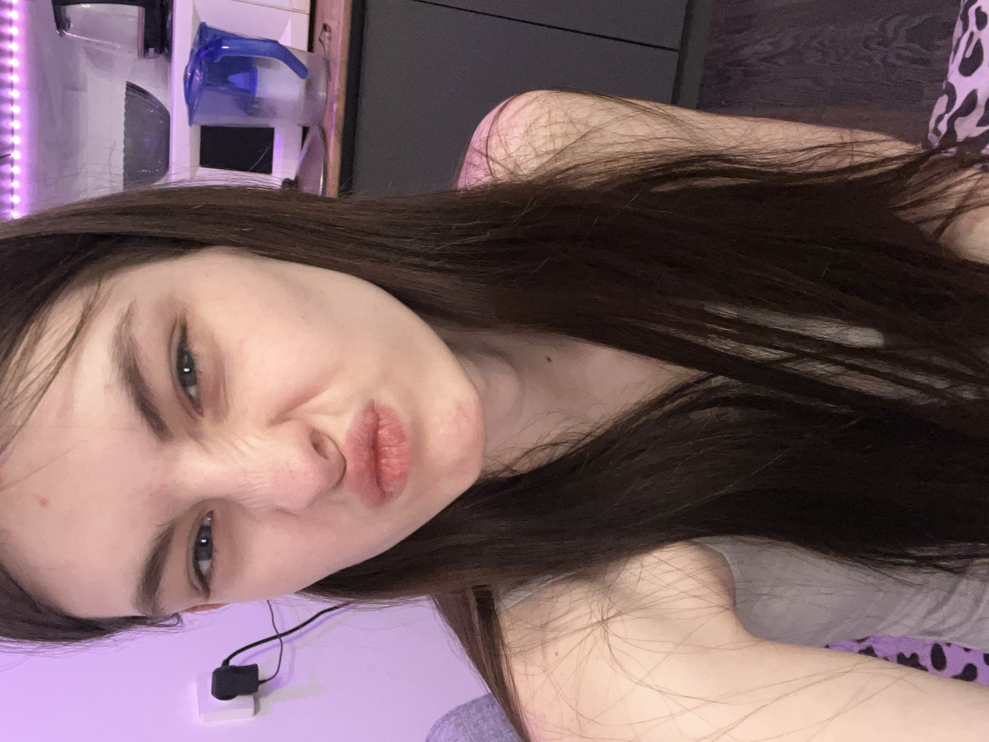 A selfie portrait of a woman with long, dark brown hair, looking directly at the camera with pursed lips. The background is a mix of colors, including a prominent purple hue, and blurred elements suggesting an interior space. There is a white object with a black plug and cable near the bottom left corner. In the upper part of the image, there's a glimpse of household items and possibly furniture, hinting at a domestic setting. The location cannot be determined from the image and any location stated would be speculation.