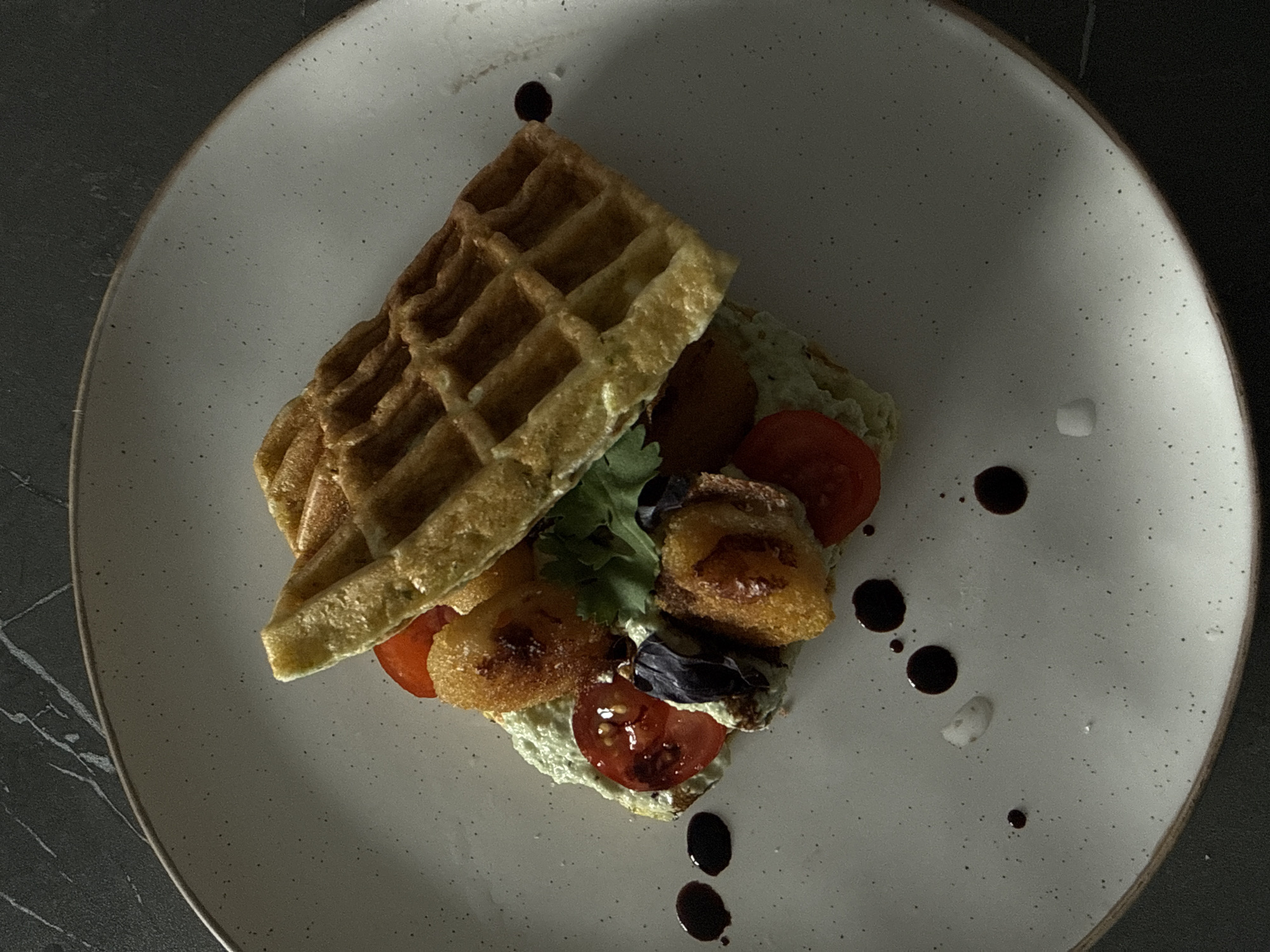 The image depicts a plate of food. The food consists of a waffle sandwich with a green spread, sliced tomatoes, and fried pieces of food. A small amount of greenery is visible. The plate is decorated with drops of dark sauce and white sauce. The scene is shot from above. The background is dark with a marble-like pattern. The plate appears to be in a restaurant. There is no indication in the picture that this image was taken in Krasnoyarsk, Russia.