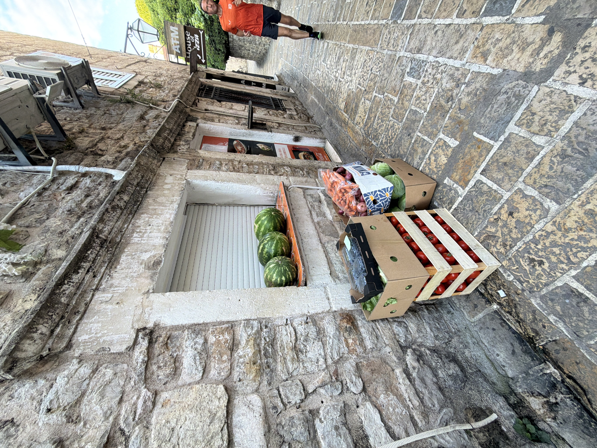 In Budva, Montenegro, a narrow cobblestone street is lined with stone buildings. On the left, a weathered stone wall features a recessed window with a white roll-down shutter. In front of the window, a ledge displays four watermelons. Below the ledge, stacked boxes contain produce: a wooden crate filled with tomatoes, and cardboard boxes with lettuce and blueberries. Further down the street, a man in an orange t-shirt and dark shorts runs towards the viewer. He is on the right side of the street, a short distance from an ATM sign and a "House of Pizza" sign. The street is illuminated by a street lamp.