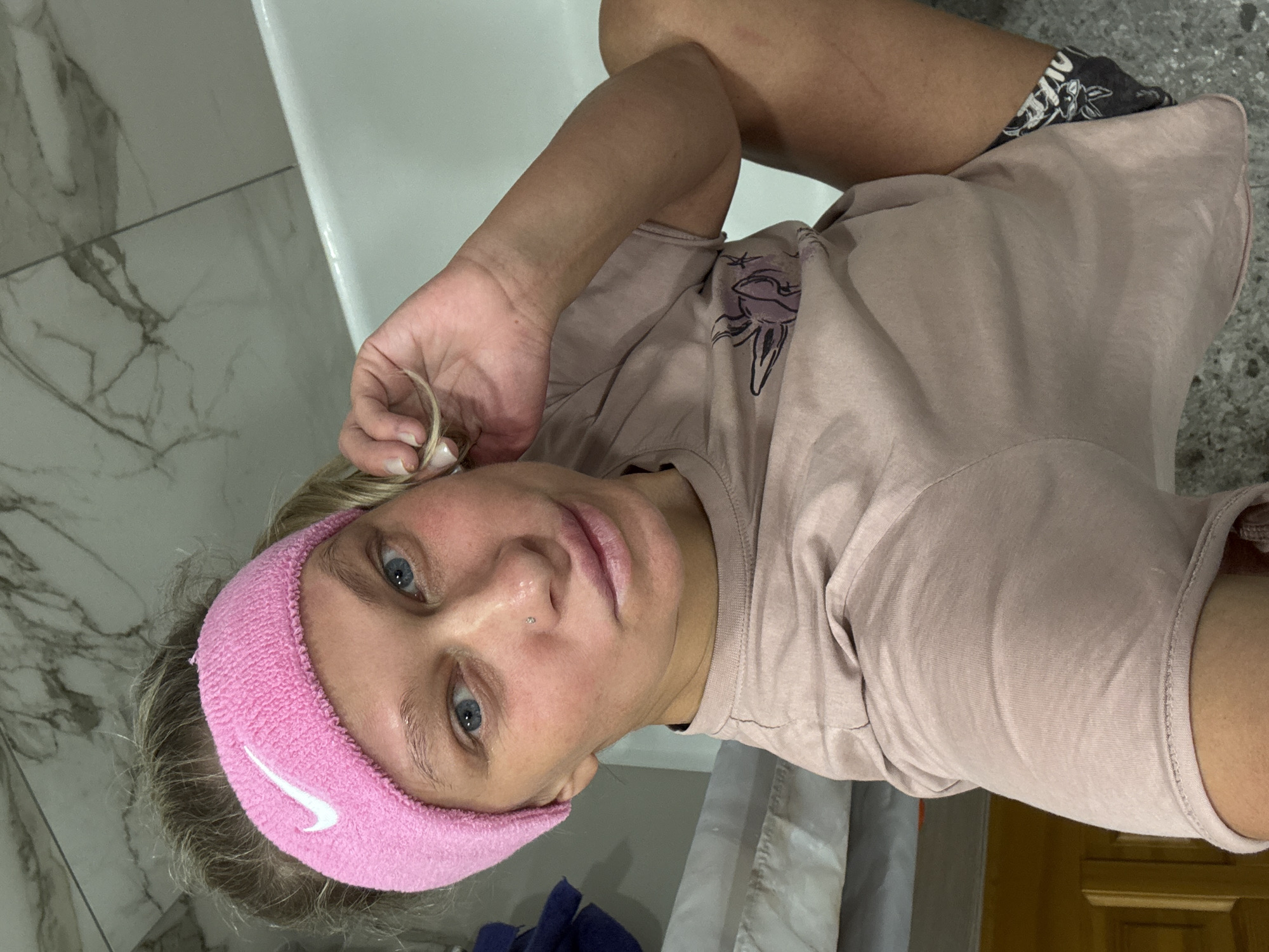 A woman with blond hair and blue eyes, wearing a pink Nike headband and a light pink t-shirt with a black graphic, is pictured in what appears to be a bathroom. She is holding a strand of her hair near her ear. The bathroom has white tiling and a bathtub. The location is likely not in the city of Perm, Russia, as no elements indicate a specific geographic location.