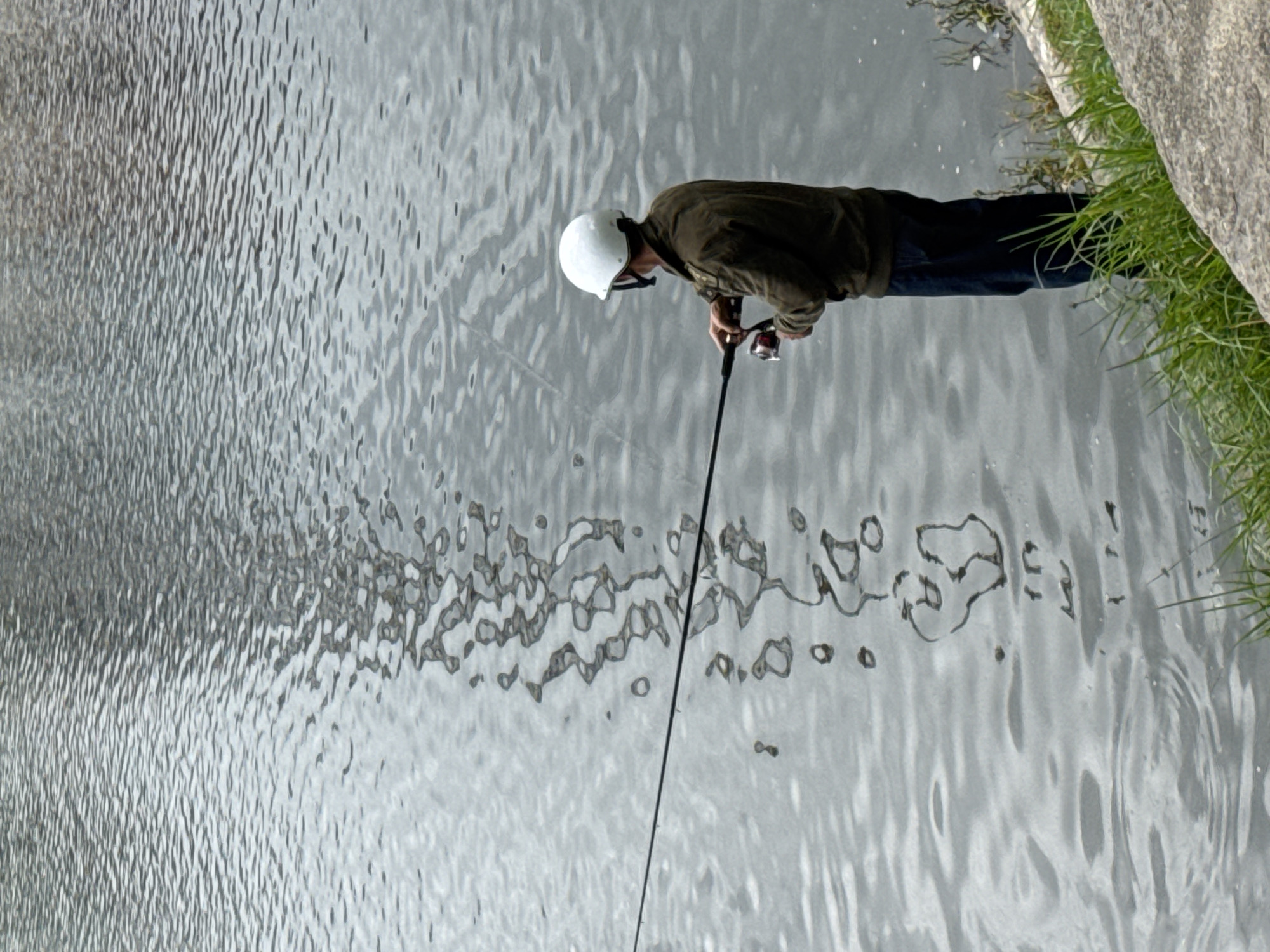 A person fishes in a body of water, likely a river or lake, wearing a white helmet and a dark jacket. The person is holding a fishing rod with a reel. The water's surface is rippled and reflects light. The background is out of focus, showing a textured surface. The foreground features green grass and a rocky bank. The image appears to be taken in Da Lat, Vietnam, a city known for its natural beauty and water bodies.