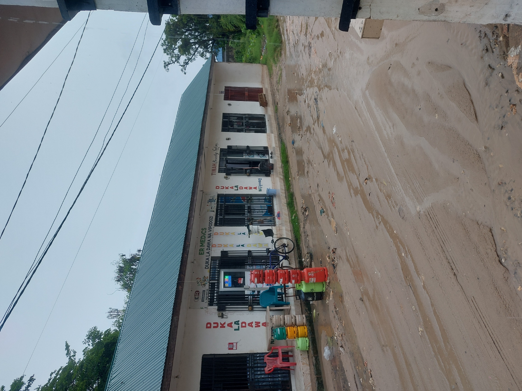 The image shows a street scene in Dar es-Salaam, Tanzania. The ground is wet and appears muddy. A building occupies the left side of the frame. Signs on the building advertise a beauty salon named "TINAH Beauty Salon" and a medical business called "ER MEDICS" which sells medicines and cosmetics ("DUKA LA DAWA NA VIPODOZI"). Cylindrical tanks, some red and some green, are stacked in front of the building along with green and yellow containers, and a red plastic chair is also present. A blue bicycle is parked against the building's exterior.