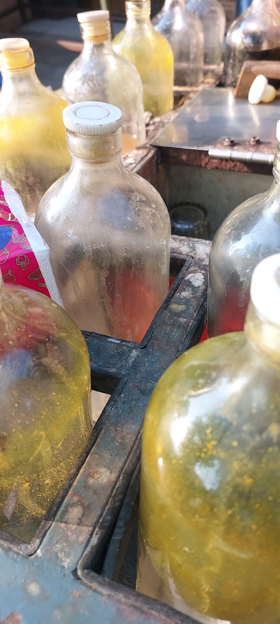 A collection of glass bottles, some filled with yellow and red liquids, are arranged in a metal container on a table. The bottles have various caps, including white plastic ones. The scene appears to be outdoors, possibly at a market stall. The overall impression is one of a roadside vendor selling beverages. The context of Semarang, Indonesia, suggests that this might be a local drink stand, possibly selling traditional Indonesian drinks like jamu.