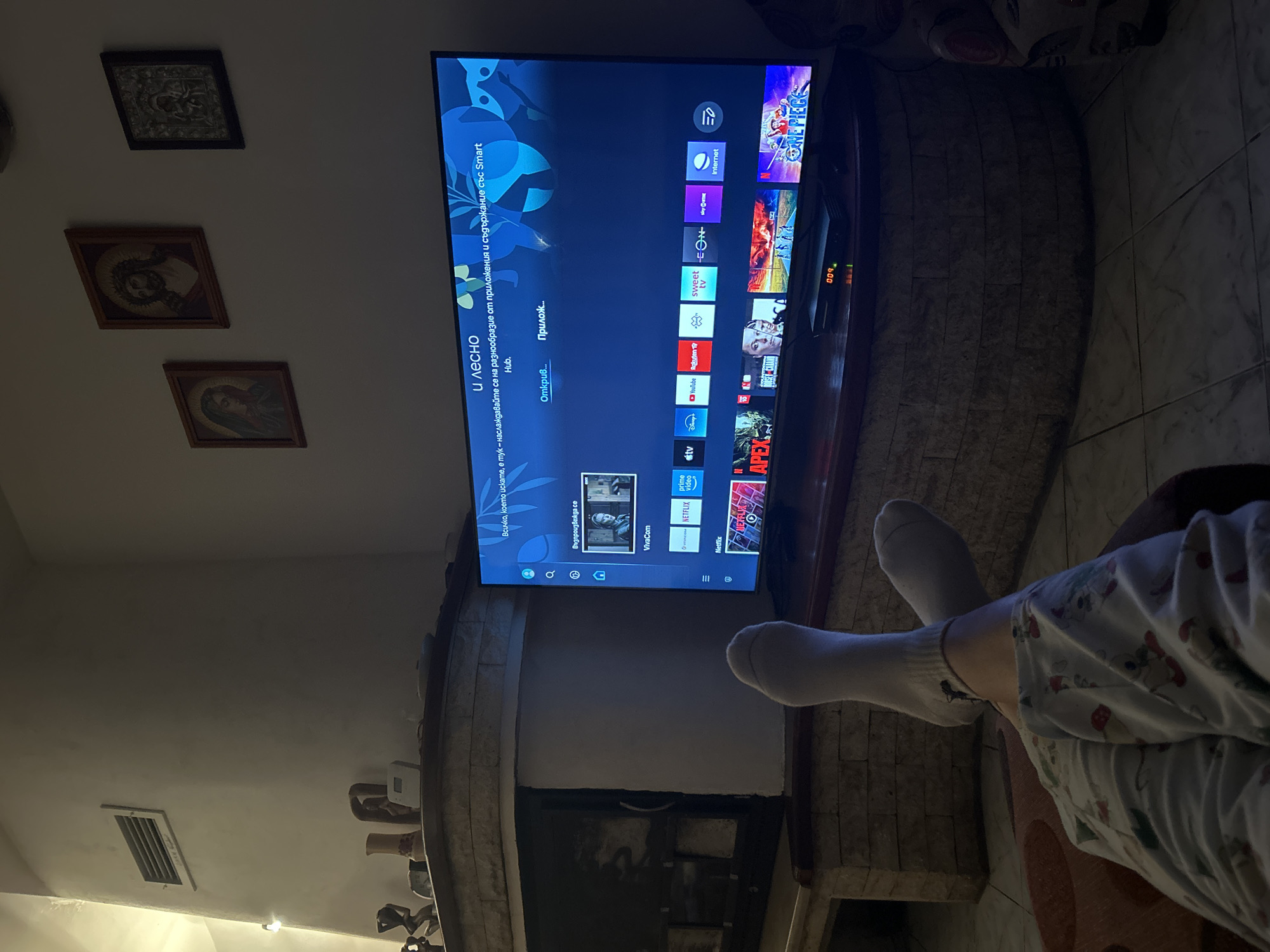 The image depicts a person sitting and watching television. The television is mounted above a fireplace, and the person's legs are visible, crossed and with white socks on. The TV displays a smart hub screen with various streaming apps and options in Bulgarian. The background includes religious icons on the wall and decorative items on the fireplace mantel. The environment appears to be a home interior. There are no specific indicators of Dolna Dikanya, Bulgaria, beyond the Bulgarian language on the television screen.
