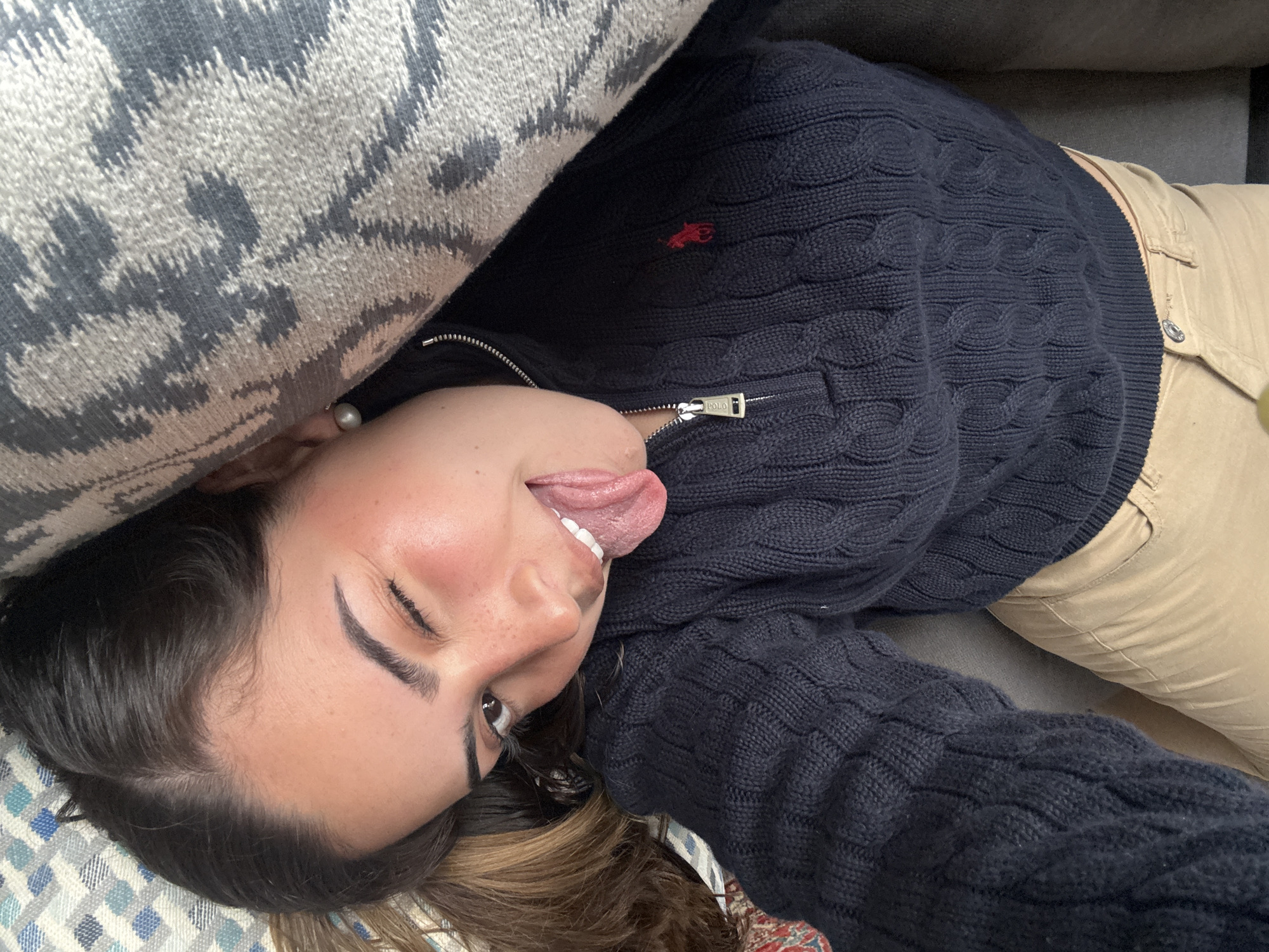 A person with dark hair is lying on a surface with patterned fabric. The person is wearing a navy blue cable knit sweater with a red logo and light tan pants. They are sticking their tongue out. A patterned gray and white pillow is near their head. The location is not discernible.