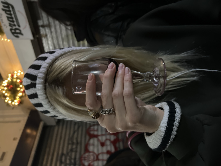 A person with blonde hair and a striped beanie is holding a wine glass. They wear a dark jacket with a white and black striped cuff. Rings are visible on their fingers, including one shaped like a fish with the word "JESUS" inscribed on it. Christmas lights are blurred in the background, along with a sign that says "Brady."