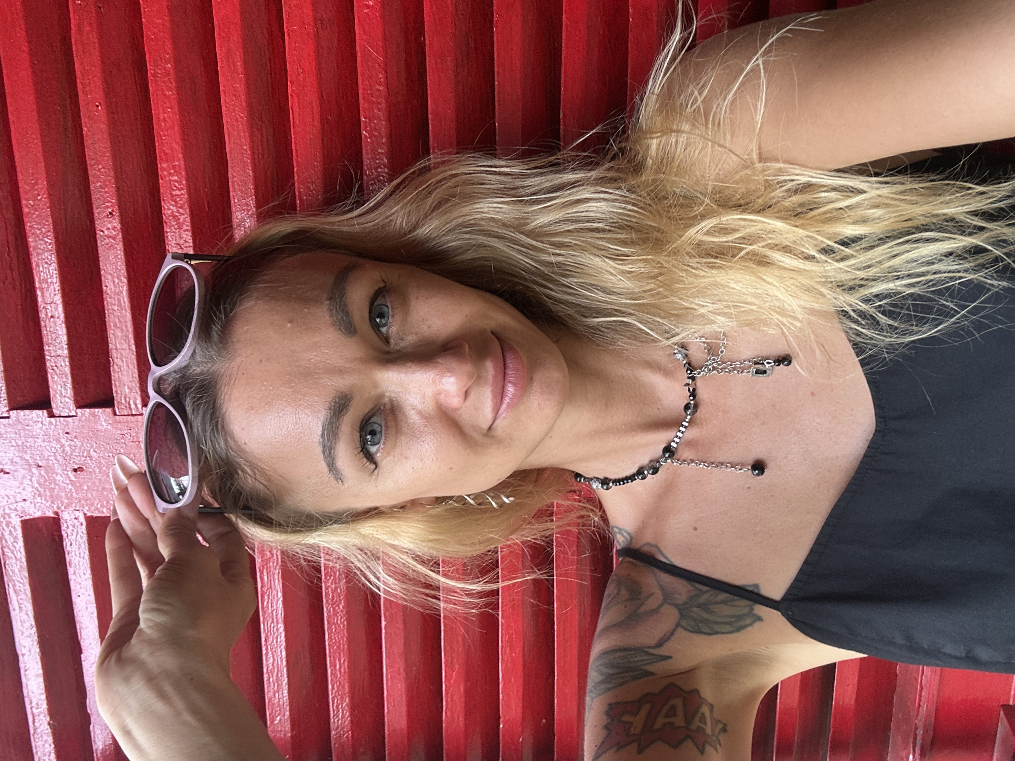 The image is a medium shot of a blonde woman in Mũi Né, Vietnam, against a red wooden shuttered background. The woman is wearing a black tank top and a beaded necklace. She is holding a pair of light purple sunglasses on top of her head with her right hand. Her left arm, visible on the left side of the image, has several tattoos. The lighting suggests it is daytime.
