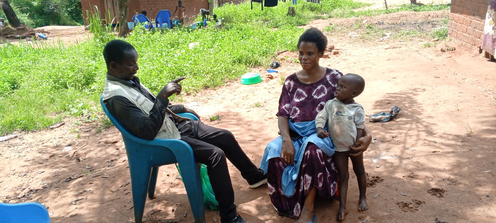 A medium outdoor shot depicts a man, a woman, and a child in Makungu, Democratic Republic of the Congo. The man sits on a blue plastic chair, dressed in a black shirt, dark pants, and a light-colored vest. He gestures with his right hand. The woman sits on a similar blue plastic chair, holding a young child. She wears a purple and white tie-dye dress. The child wears a gray shirt. The ground is dirt and shadows, with some green foliage in the background. Other people and blue plastic chairs are visible in the distance. A green bowl sits on the ground behind the woman. A black plastic bag is near the woman and child. The edge of a brick building is visible to the right.