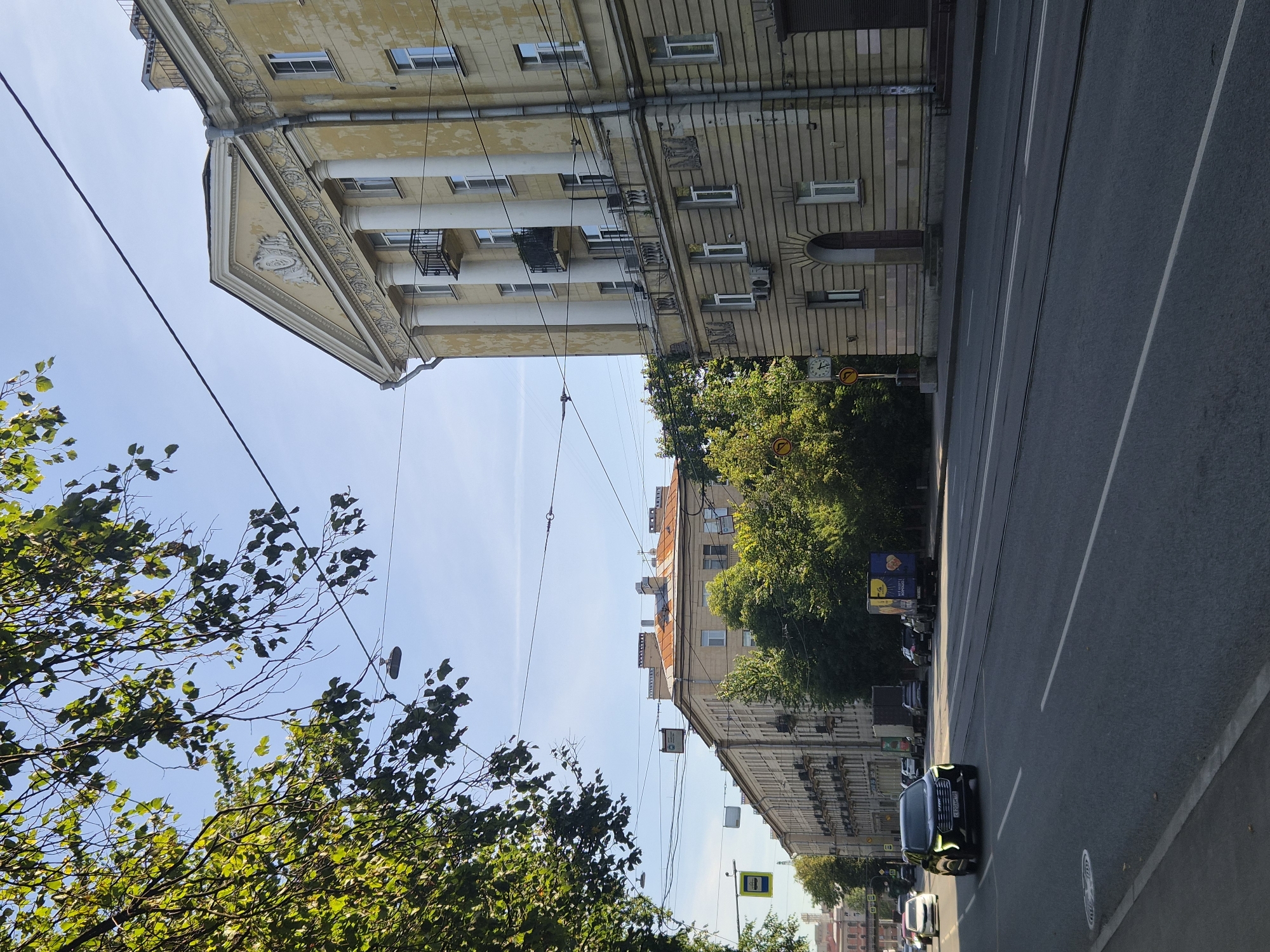 The image depicts a street scene in Saint Petersburg, Russia. In the foreground, a multi-lane asphalt road stretches into the distance. A black car is driving in the left lane of the road. On the right side of the road, a large, light-yellow building with classical architectural elements, including columns and a triangular pediment, stands prominently. Adjacent to this building, further down the street, is another long, light-colored building with a rusty-orange roof. Trees with green foliage are visible along the sidewalk and between the buildings. Overhead, a network of power lines crisscrosses the sky, which is a clear blue with a few thin clouds. The overall scene suggests an urban environment on a bright, sunny day.