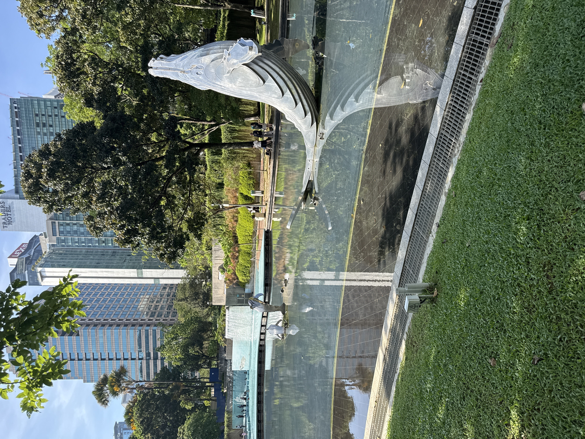 The image shows a silver whale sculpture reflected in a shallow pool of water. The sculpture is positioned in a park-like setting with green grass and trees surrounding the pool. In the background, modern high-rise buildings are visible. The image is likely taken in Kuala Lumpur, Malaysia, based on the architectural style and the overall urban landscape. There appear to be people standing or walking near the far side of the pool. A metal grate borders the pool on one side.