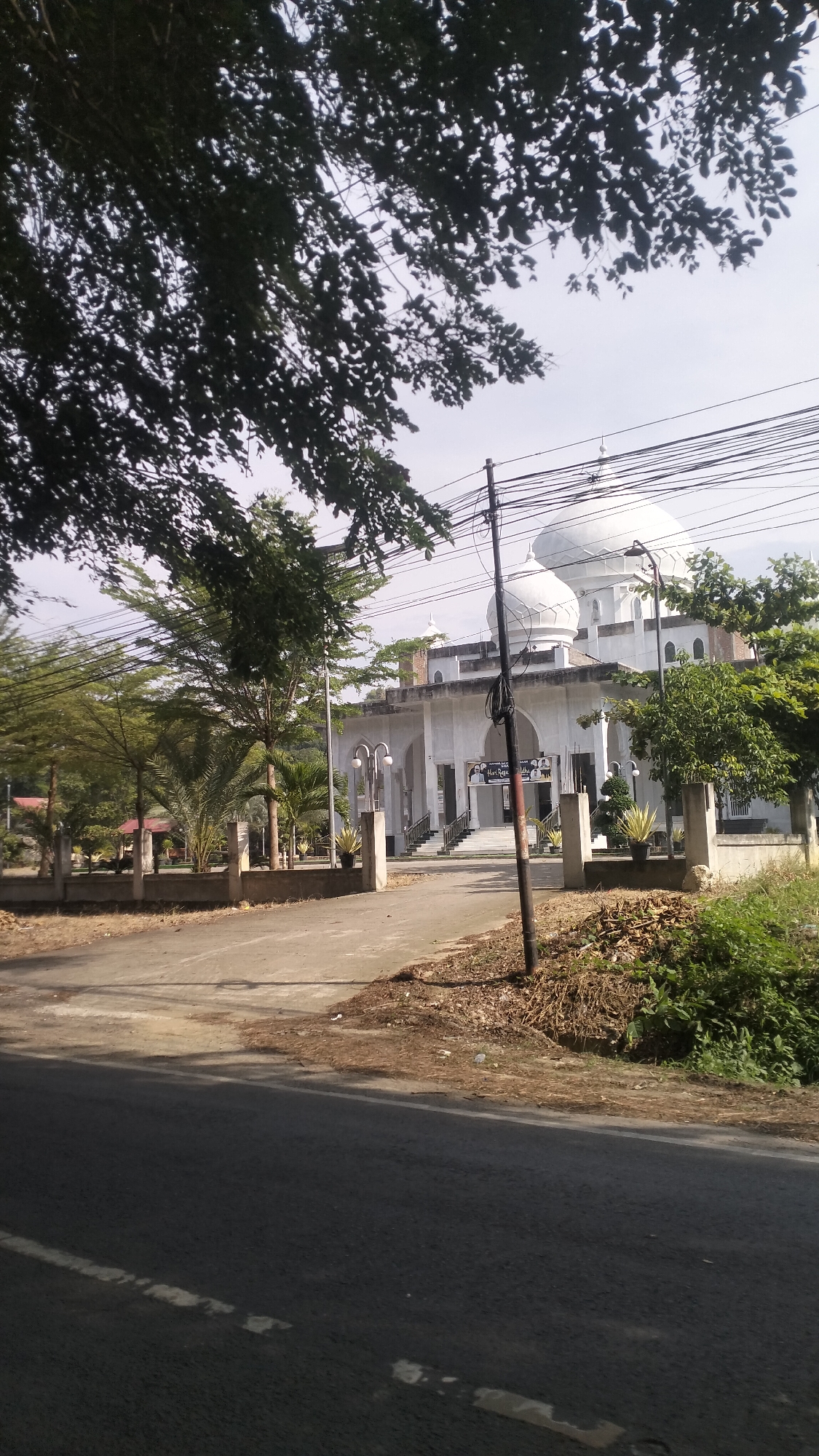 The image depicts the exterior of a mosque in Lhokseumawe, Indonesia. The mosque features prominent white domes and a white facade. The foreground shows a paved road with a white line marking. To the left of the road, there is a low concrete wall and an area with dry grass and scattered leaves. Several trees with green foliage are visible, some with a structured appearance and others more natural. In the middle ground, a pathway leads towards the mosque entrance, which has stairs and is flanked by decorative lamps. A utility pole with numerous overhead wires stands to the right of the pathway, in front of the mosque. The background consists of a bright, overcast sky and more trees. The overall scene suggests a daytime view of a religious building and its surroundings in an urban or suburban setting.