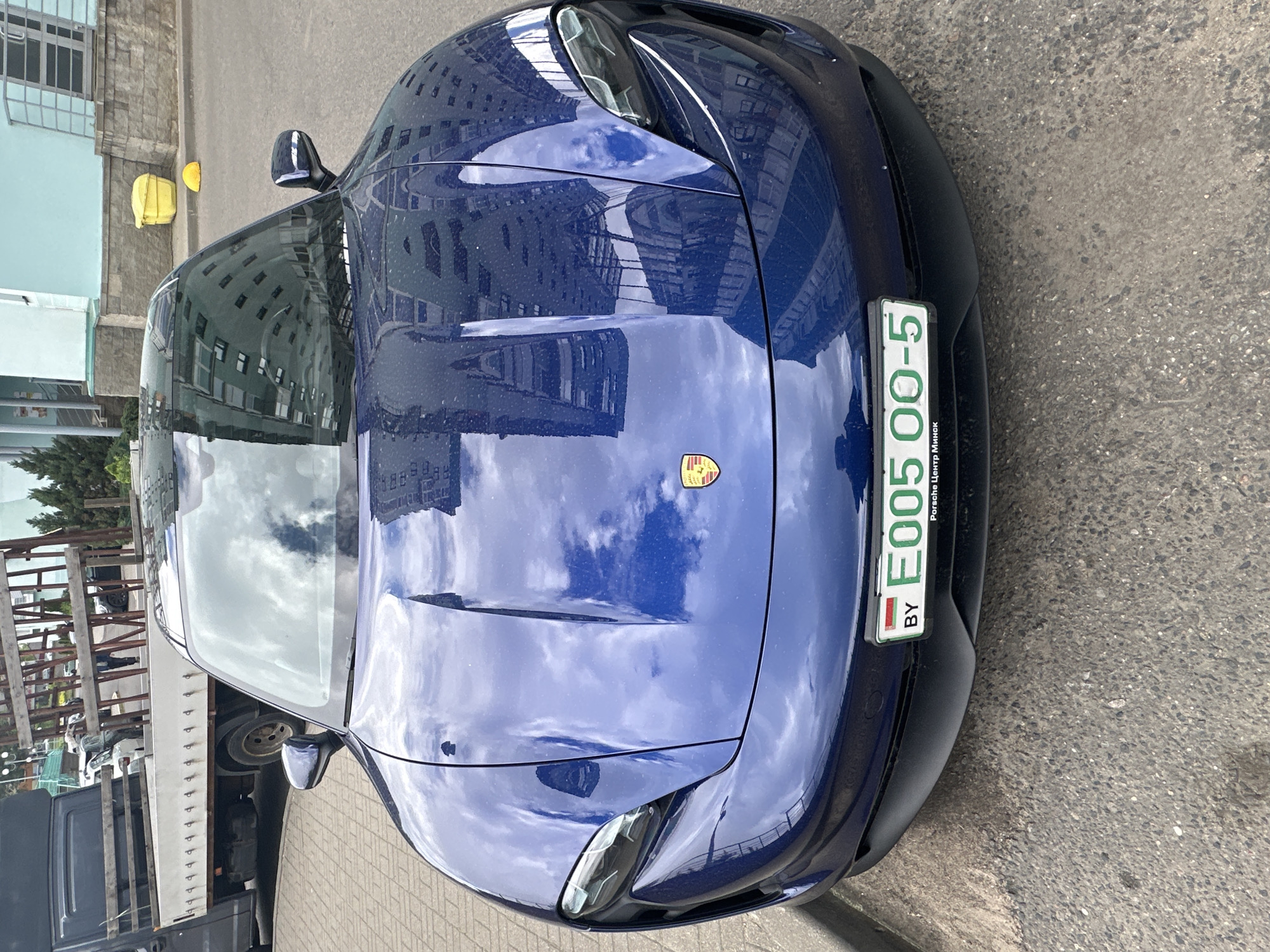 The image shows the front of a blue Porsche car with Belarusian license plate E005 00-5, indicating it is registered in Minsk. The car is parked on a paved surface in an urban area. Reflections in the car's paint suggest tall buildings nearby. There are traces of water droplets on the car. The license plate frame states “Porsche Centre Minsk.” A yellow hard hat is partially visible behind the car. The surrounding buildings have light blue or gray facades. A truck is parked behind the car on the left side.