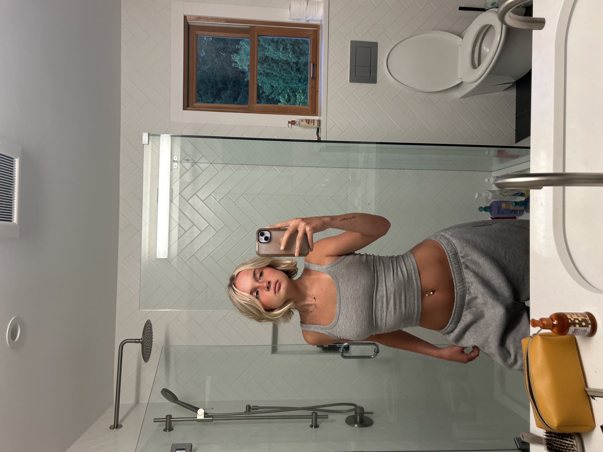 A young Caucasian female with blonde hair and rosy cheeks is taking a mirror selfie in a modern bathroom. She is wearing a grey tank top and grey sweatpants, with a belly button piercing visible. The bathroom features white herringbone tile walls, a glass-enclosed shower with a rain showerhead, a wooden-framed window with a view of trees, a white toilet with an open lid, and a white sink with a chrome faucet. On the counter, a yellow makeup bag, a bottle of hair oil, and a hairbrush are visible. The image does not contain any information relating to Squamish, Canada.