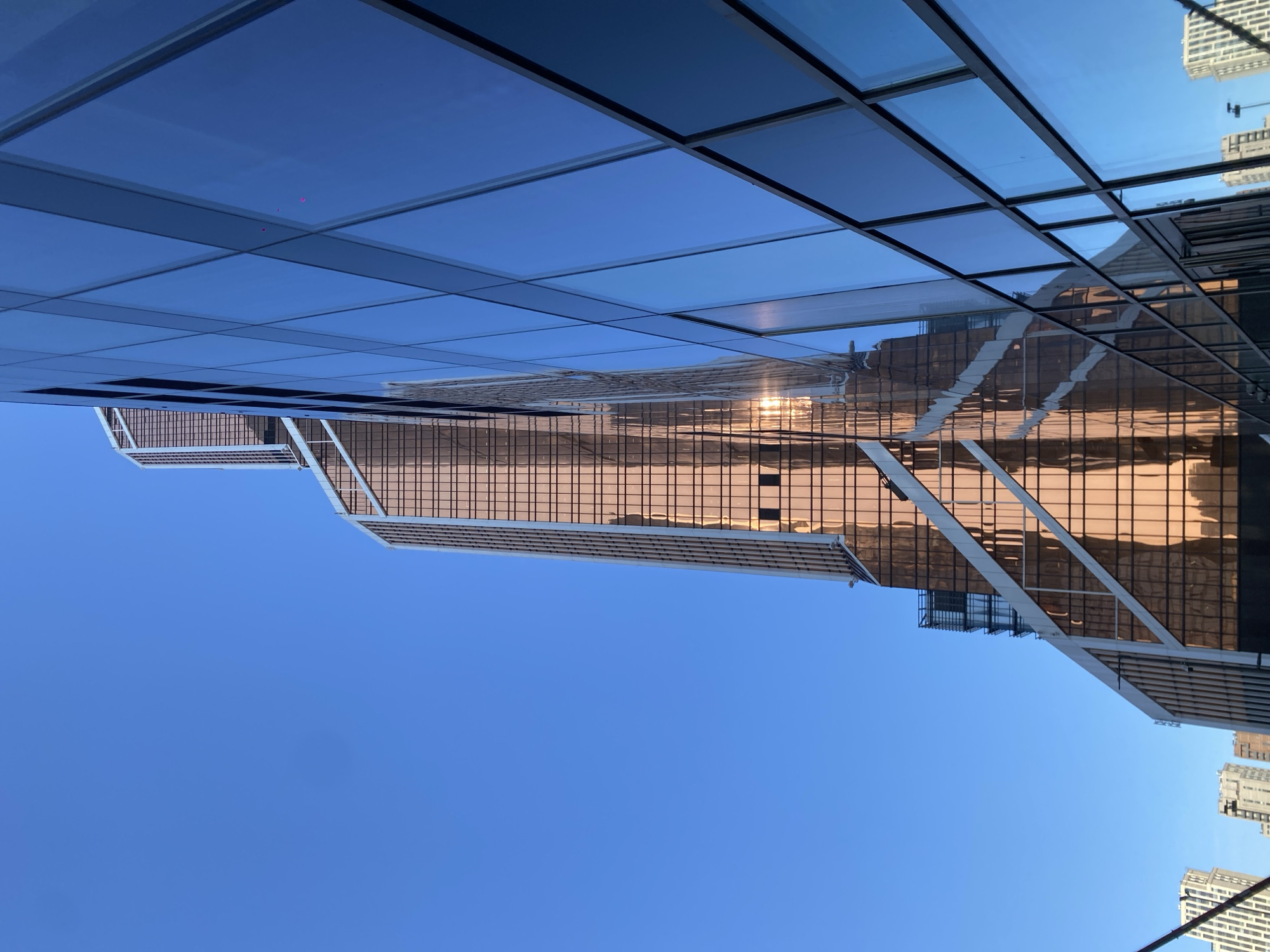 The image depicts a view looking up at a building with reflective glass facades in Khimki, Russia. The sky is a clear blue. A different building with a grid-like pattern is reflected in the glass of the closer building. Part of a glass canopy or overhang is visible in the upper portion of the frame.